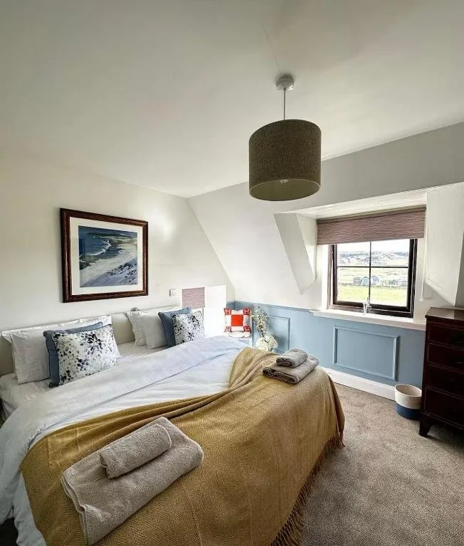 A cozy bedroom with a large bed, beige blanket, and decorative pillows, featuring a wall painting, window with blinds, and ceiling light fixture.