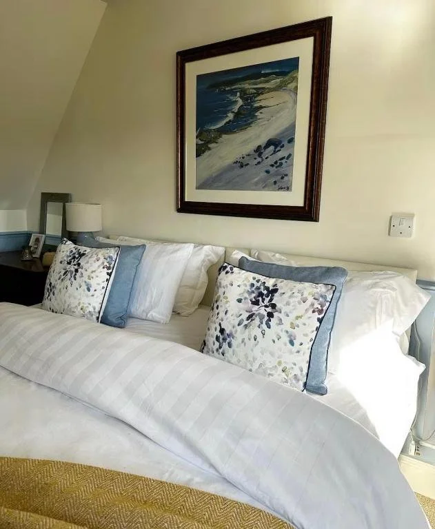 A neatly made bed with white sheets, four pillows with floral and blue pillowcases, and a framed landscape painting hanging above the bed in a cozy bedroom.