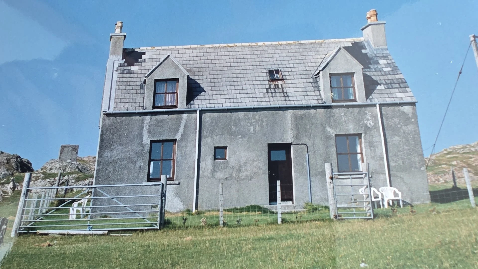 A three-story gray house with a steep roof, multiple windows, and a small fenced yard. The house shows signs of aging, with some deterioration on the roof and walls. There are white plastic chairs near the gate, and the house is set against a clear blue sky with some rocky terrain visible in the background.