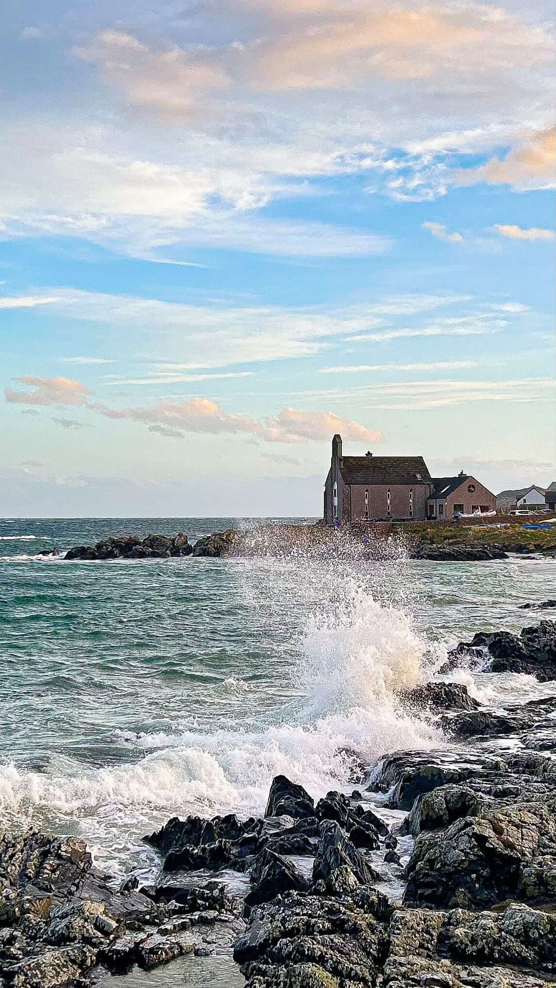 A house on a rocky shoreline with waves crashing against the rocks, under a partly cloudy sky.