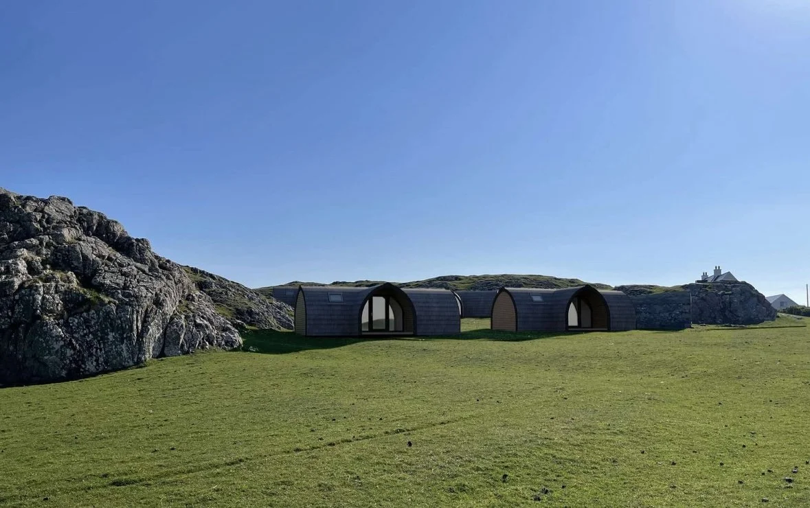 Three small modern cabins with arches in a grassy field near rocky hills and a house in the distance under a clear blue sky.