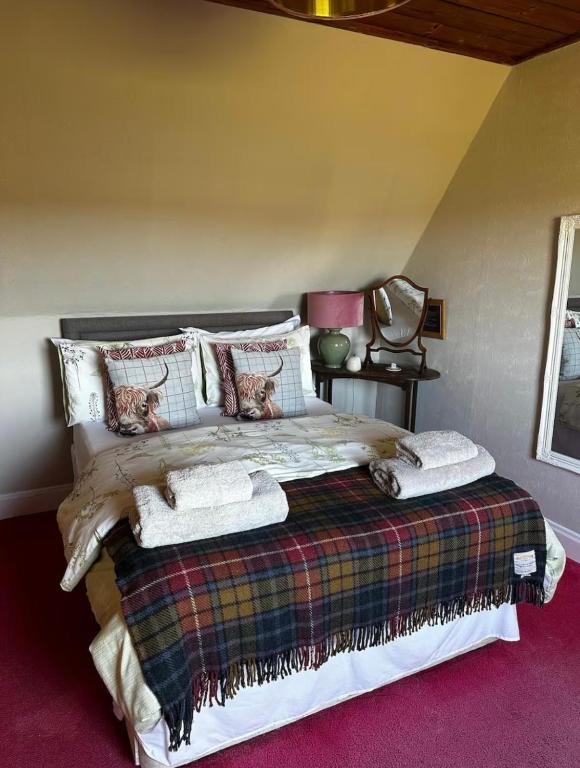 A neatly made bed with two tiger-themed pillows, covered with a plaid blanket, with two rolled towels on top. Beside the bed, on a small side table, there is a pink lamp, a green vase, and a mirror. The room has a sloped ceiling and a framed mirror o