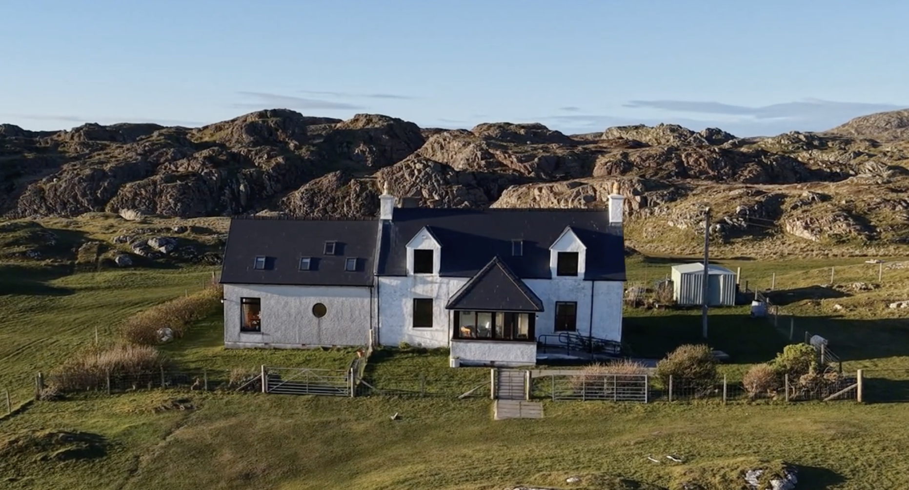 A white house with black roof shingles and multiple chimneys, located in a countryside setting with grassy lawn and a small fenced yard, with rocky hills and a blue sky in the background.