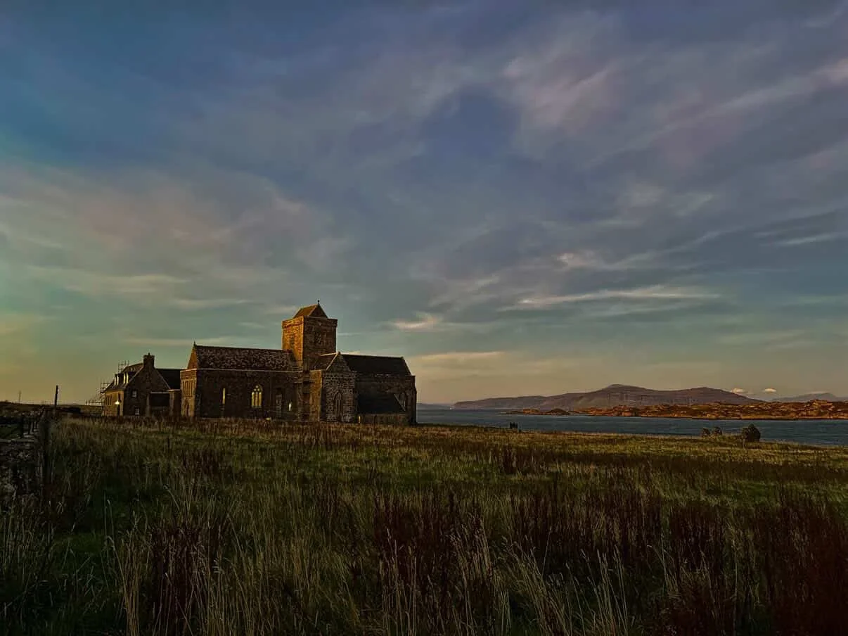 A historic stone church with a tower on the Isle of Iona - known as Iona Abbey - sits on a grassy field near a body of water, with hills in the distance and a cloudy sky overhead.