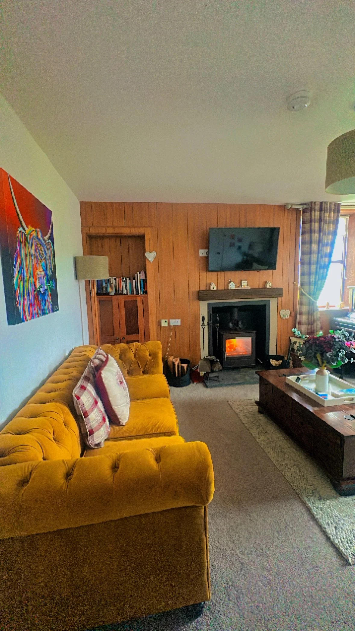 Cozy living room with yellow velvet tufted sofa, fireplace, wall-mounted TV, wooden wall paneling, window with checkered curtains, and colorful artwork.
