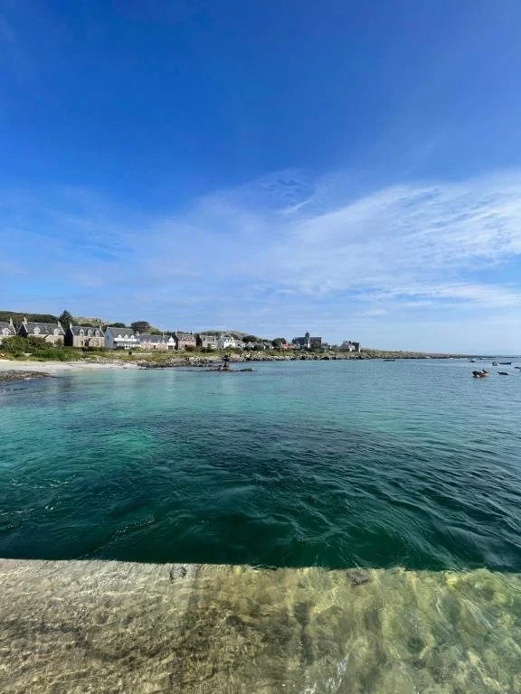 A coastal scene with clear blue water, a small beach, and houses along the shoreline under a partly cloudy sky.