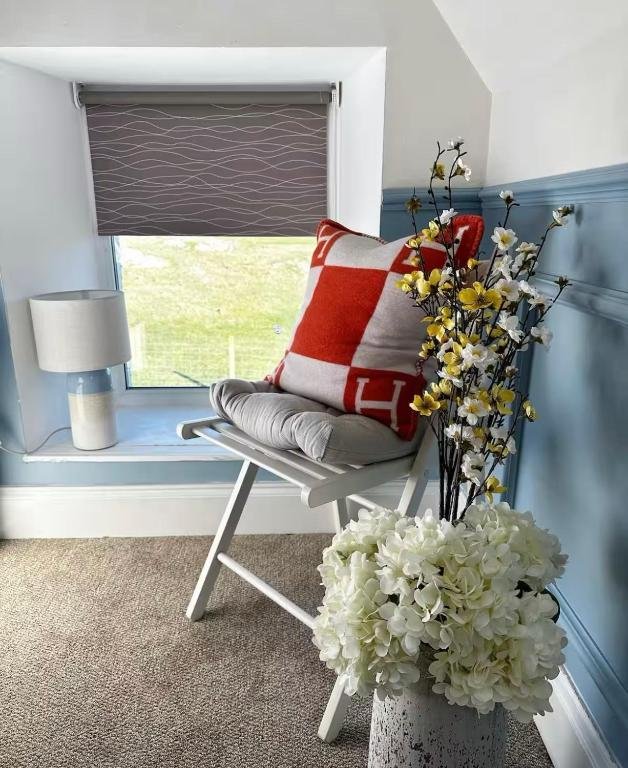 A cozy corner with a white wooden chair holding a large red and white throw pillow and a gray cushion. Next to the chair is a tall vase with yellow and white flowers, and another white vase with a lamp is on the windowsill. The window has a gray roll