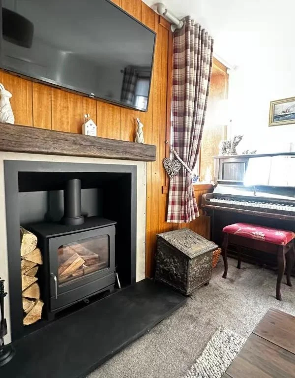 Cozy living room corner with a fireplace, wood-burning stove, and firewood stacked beside it, topped by a wooden mantle with decorative figurines. A flat-screen TV is mounted above the mantle. To the right, a plaid curtain partially covers a window, 