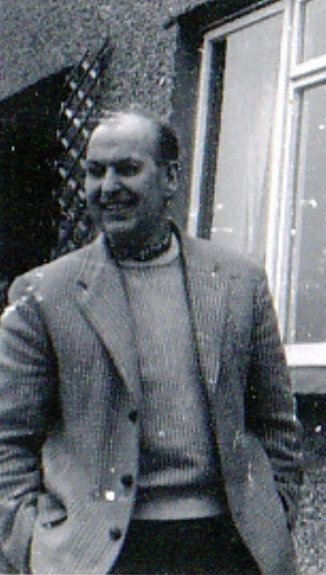 A man smiling outdoors, wearing a blazer over a sweater, standing in front of a building with a window and a trellis.