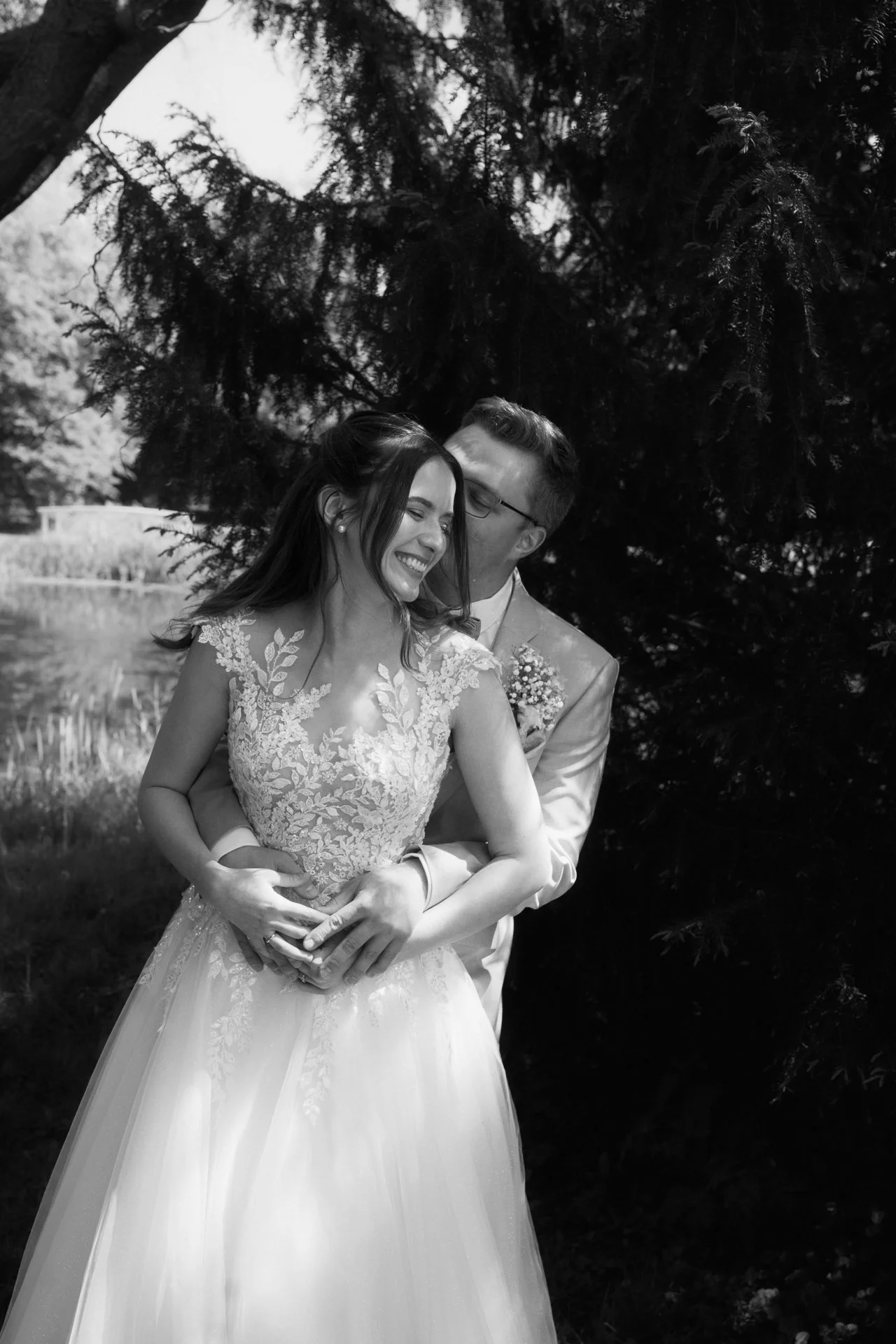 Ein glückliches Brautpaar umarmt sich im Freien, im Schatten eines Baumes, in einem Hochzeitskleid und Anzug.
