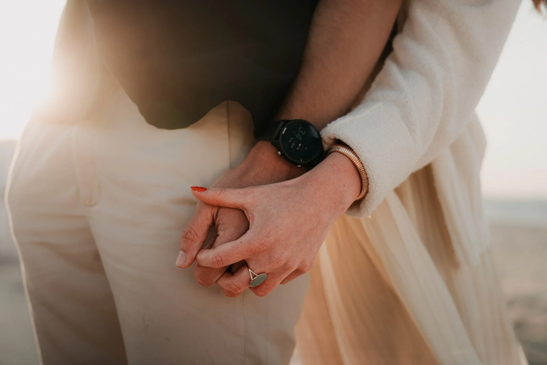 Nahaufnahme von einem Paar, das sich an der Hand hält, mit einer Person, die eine schwarze Smartwatch trägt, im Hintergrund ein Strand bei Sonnenuntergang.