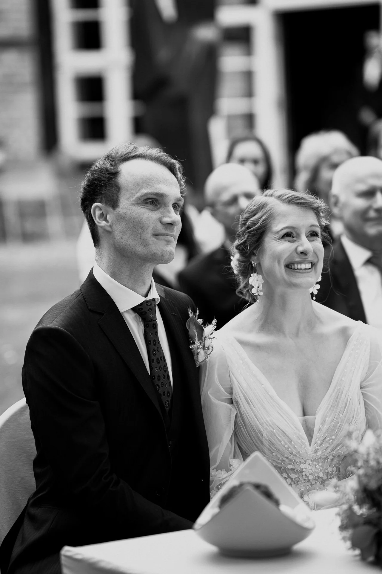 Ein Hochzeitsfoto in Schwarz-Weiß von einem glücklich lächelnden Brautpaar bei einer Zeremonie, umgeben von Gästen.