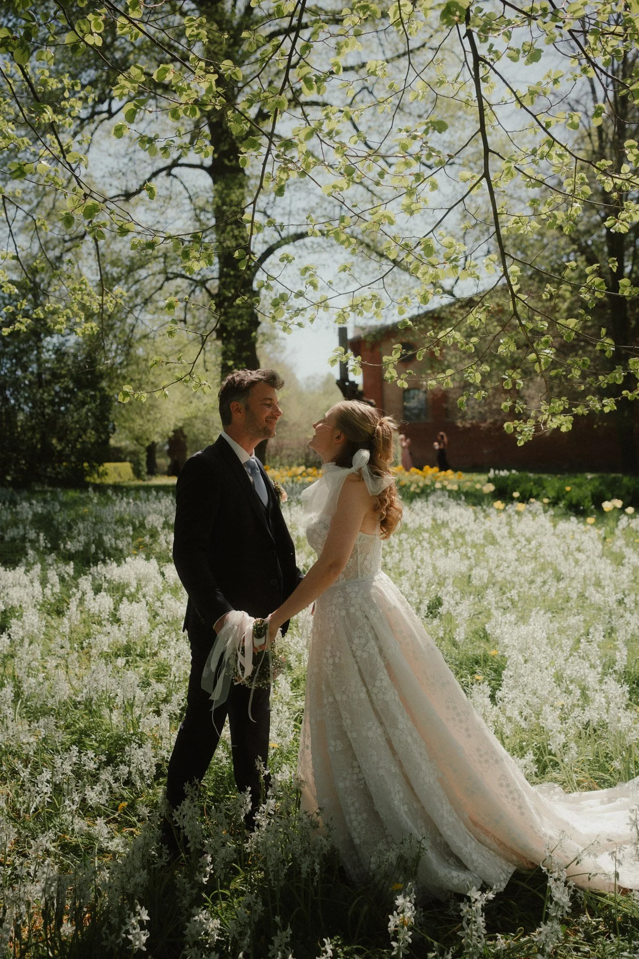 Ein Brautpaar hält sich an einem blühenden Baum in einem Garten bei Sonnenlicht. Die Frau trägt ein weißes Hochzeitskleid, der Mann einen schwarzen Anzug.