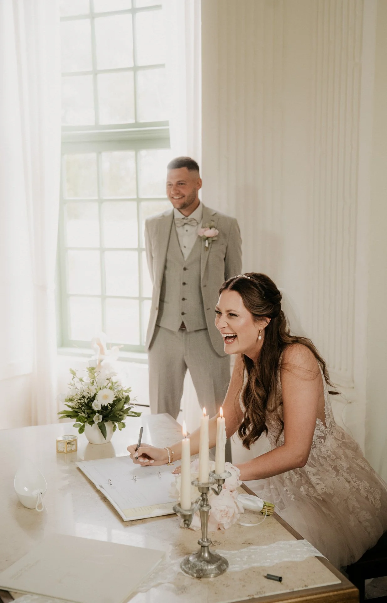 Ein Brautpaar bei einer Eheschließung. Die Braut sitzt lachend an einem Tisch und unterschreibt ein Dokument, während der Bräutigam im grauen Anzug im Hintergrund steht und lächelt.