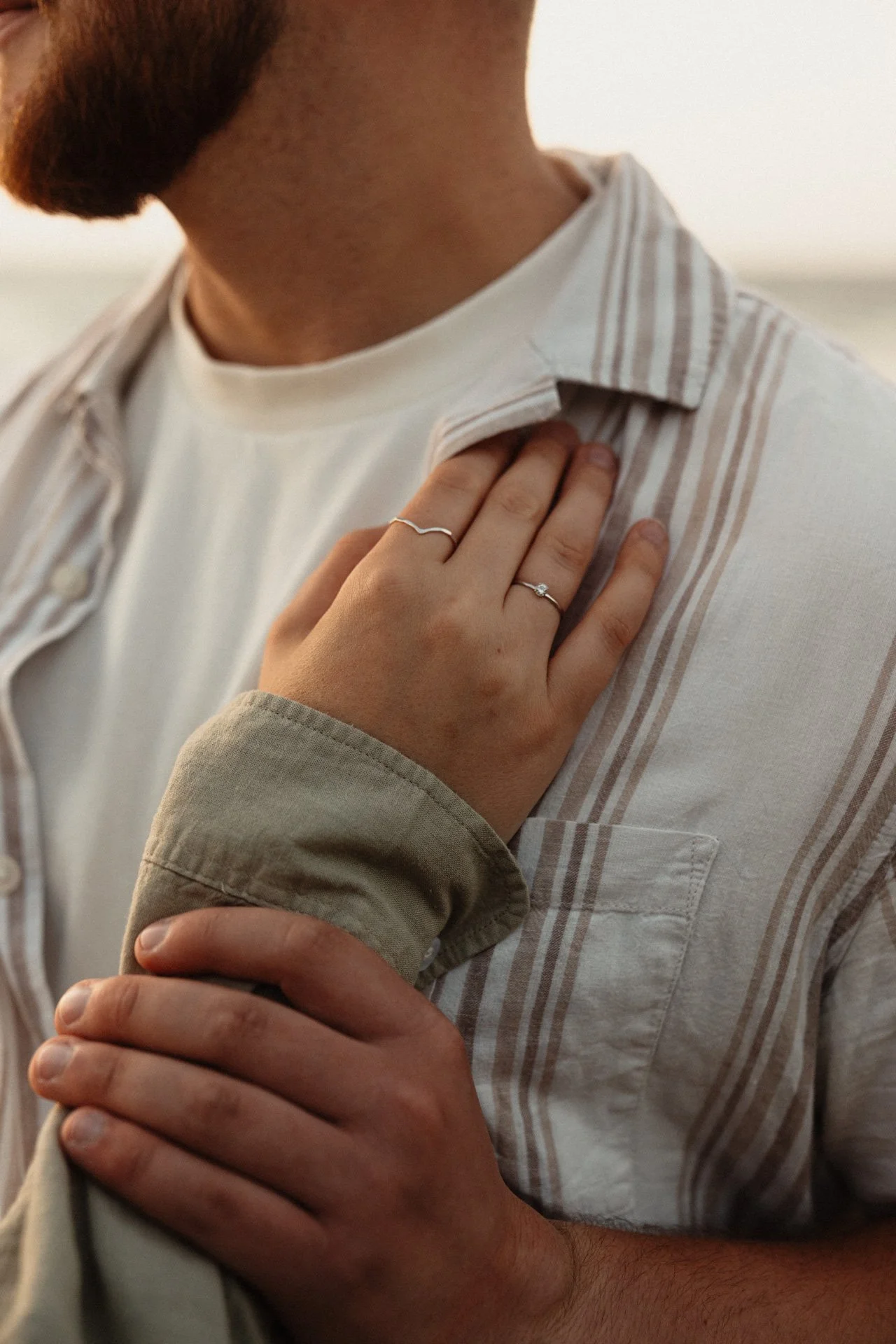 Nahaufnahme eines Mannes in einem weißen, gestreiften Hemd, die Hand auf die Brust gelegt, trägt einen Ehering und einen weiteren Ring, der Person, die den Arm hält, trägt eine braune Jacke.
