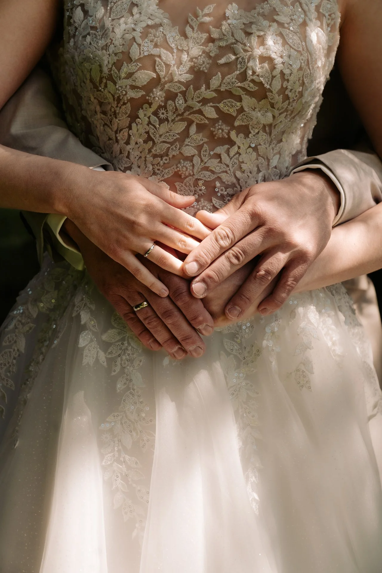 Nahaufnahme der Hände zweier Menschen, die einander in einer Umarmung halten, mit einem Hochzeitskleid im Hintergrund.