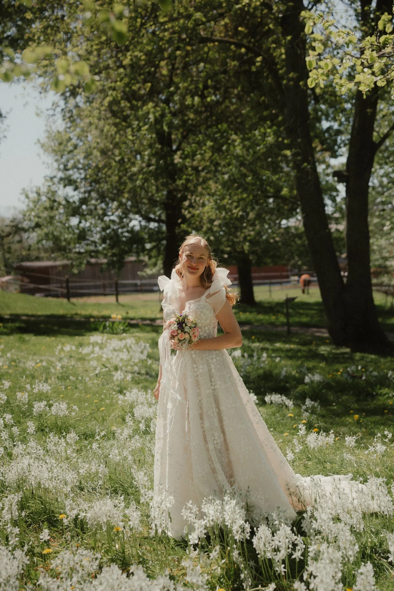 Junge Frau in Hochzeitskleid im Garten mit weißen Blumen und grünen Bäumen, lächelnd und Blumenstrauß haltend.