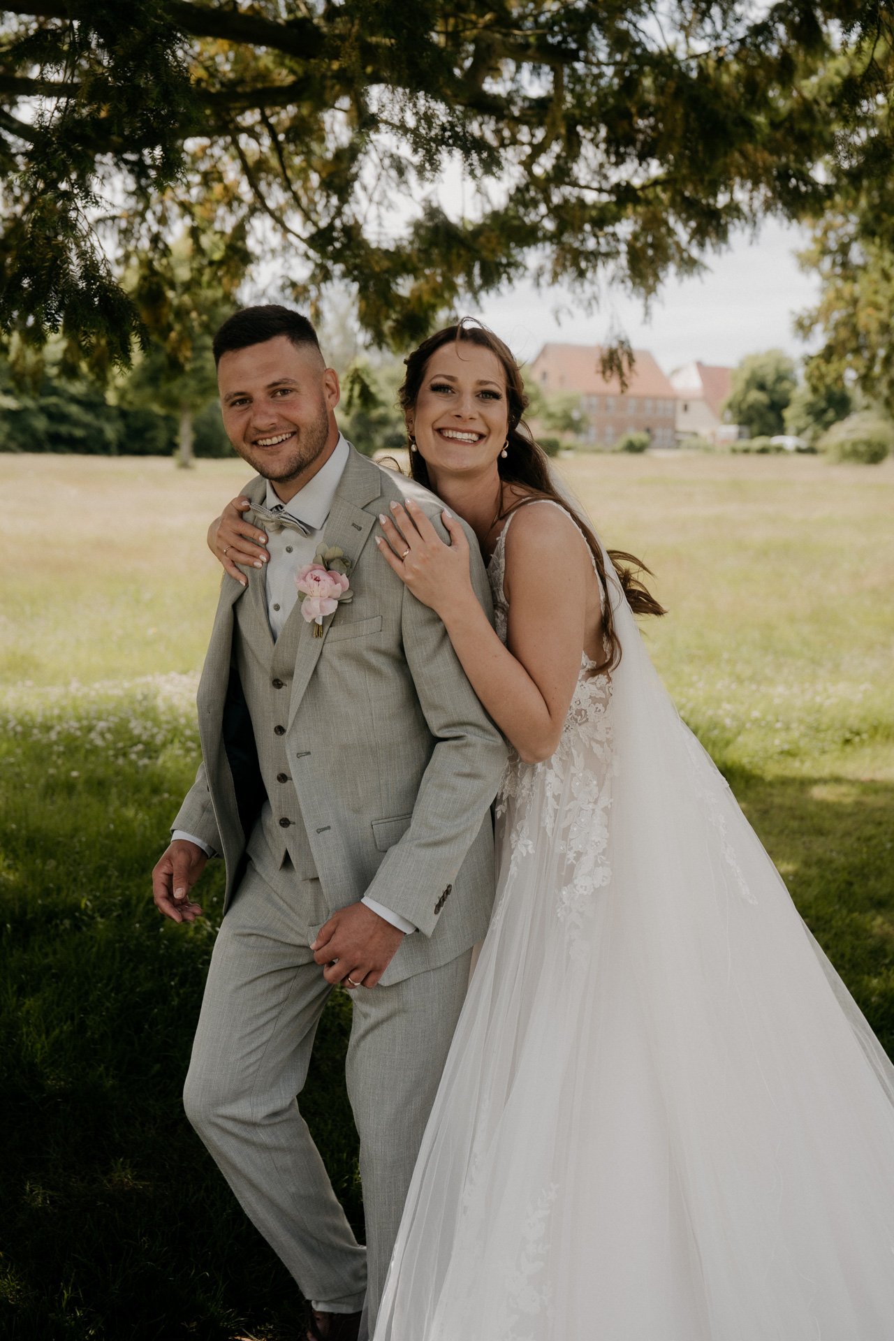 Ein glückliches Brautpaar, das im Freien unter einem Baum steht, die Braut in einem weißen Kleid und der Bräutigam in einem grauen Anzug, beide lächeln und umarmen sich.