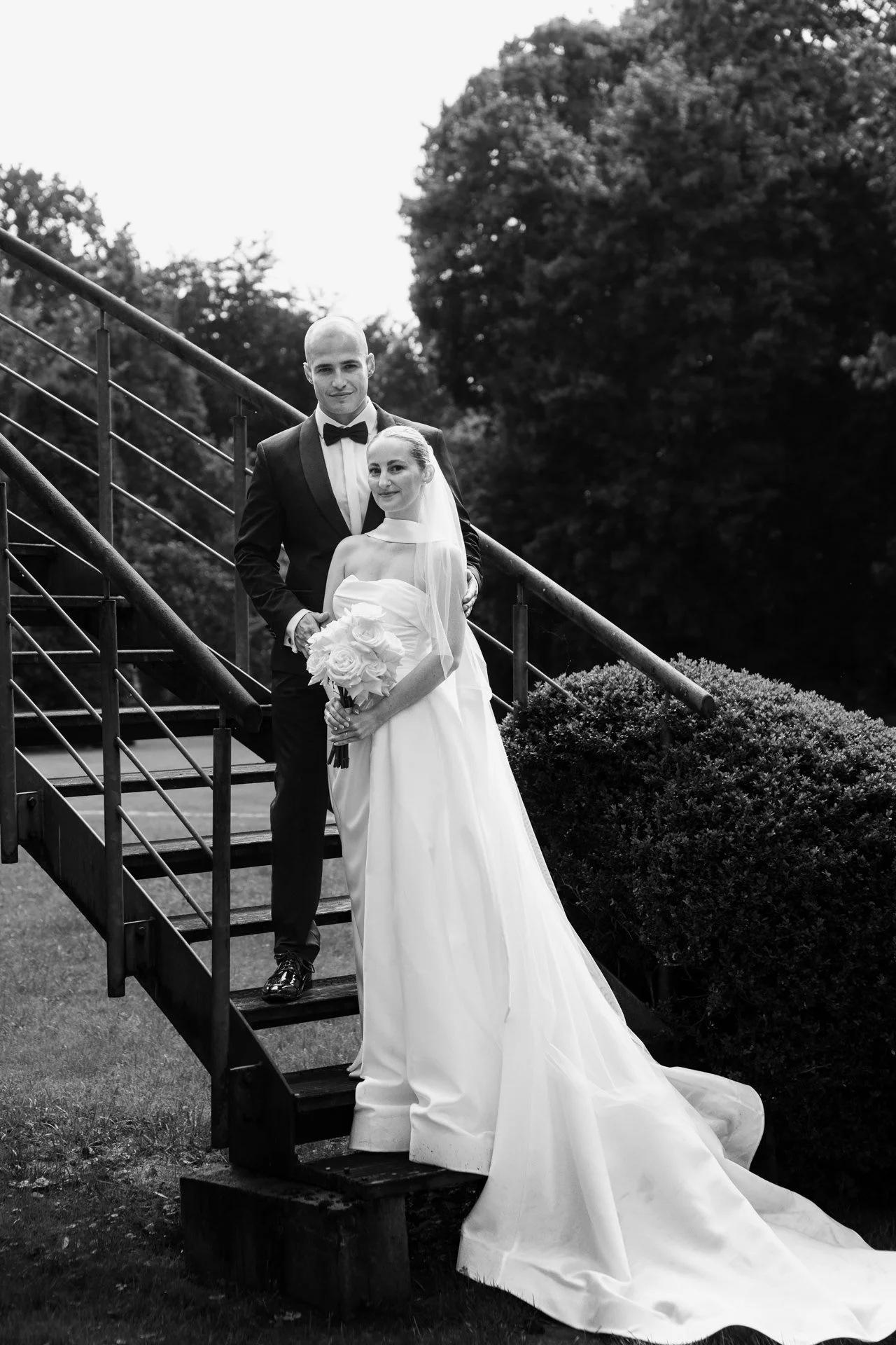 Schwarz-weiß-Foto eines Brautpaares auf einer Außentreppe, die Braut trägt ein langes weißes Brautkleid und hält einen Blumenstrauß, der Bräutigam trägt einen schwarzen Anzug mit Fliege.
