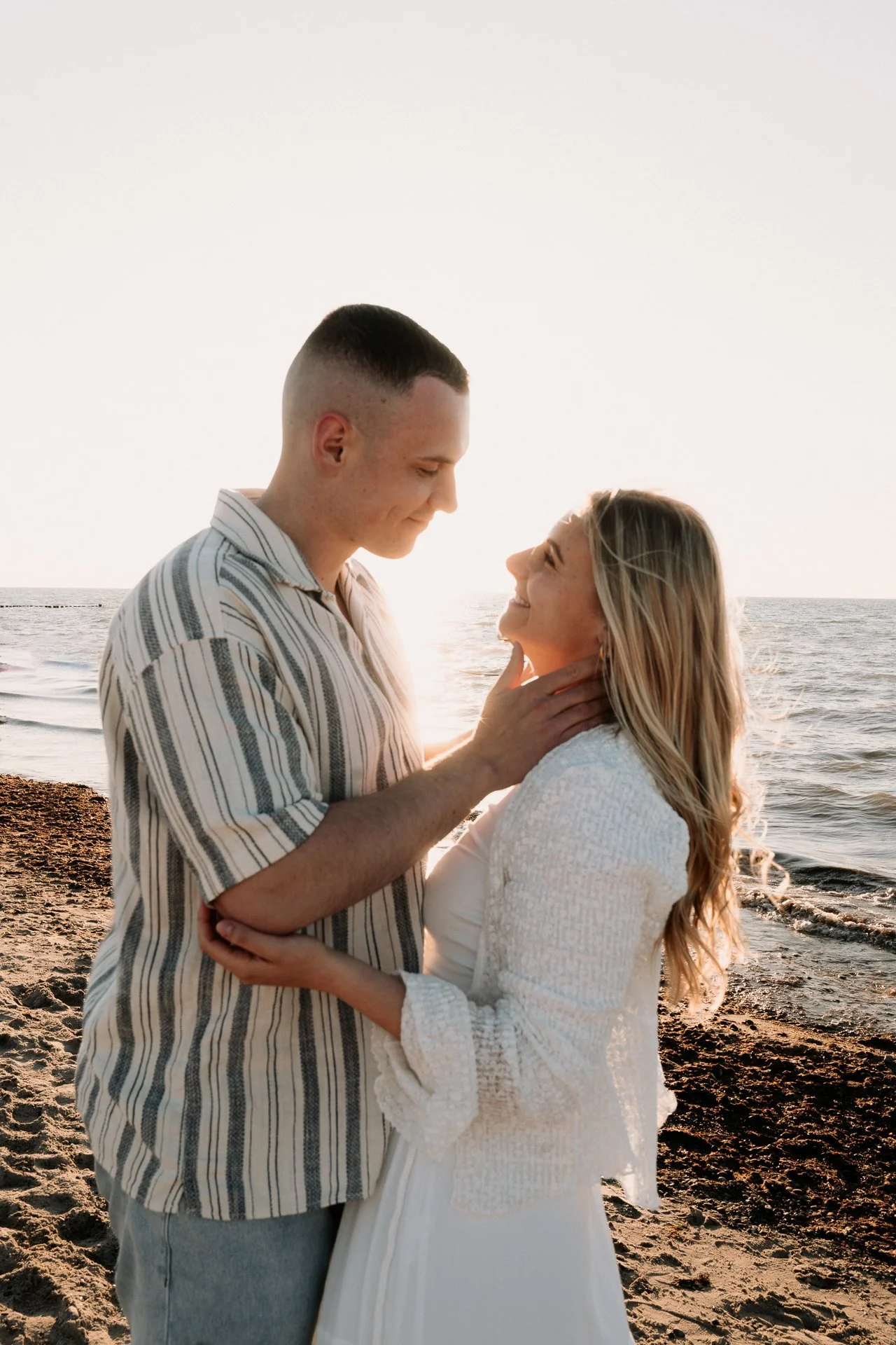 Ein verliebtes Paar steht am Strand bei Sonnenuntergang, lächelt einander an, während das Meer im Hintergrund glitzert.