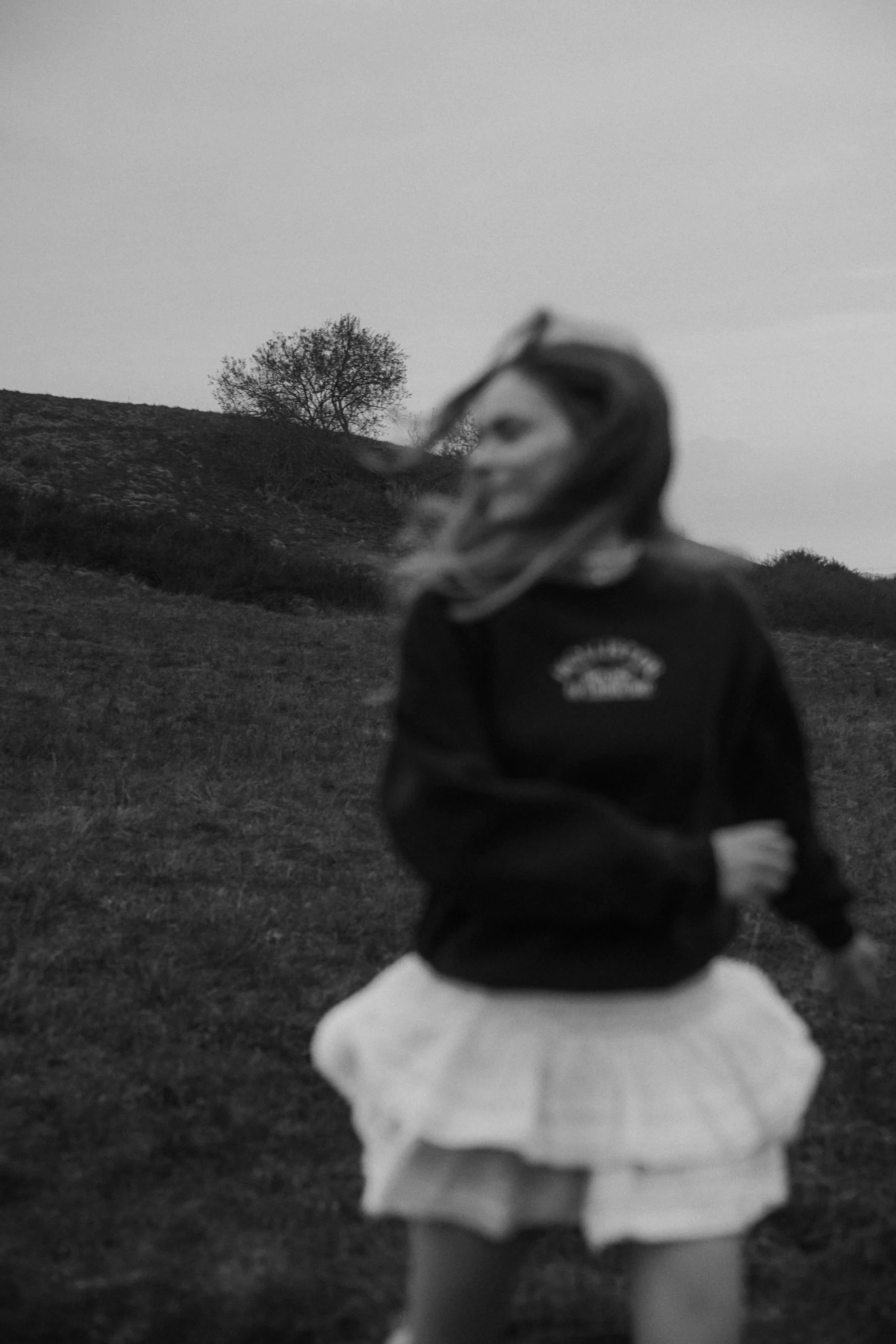 Ein Mädchen in einem schwarzen Pullover und einer weißen Rock, das auf einer Wiese steht, im Hintergrund Bäume, im Schwarz-Weiß-Foto, unscharf