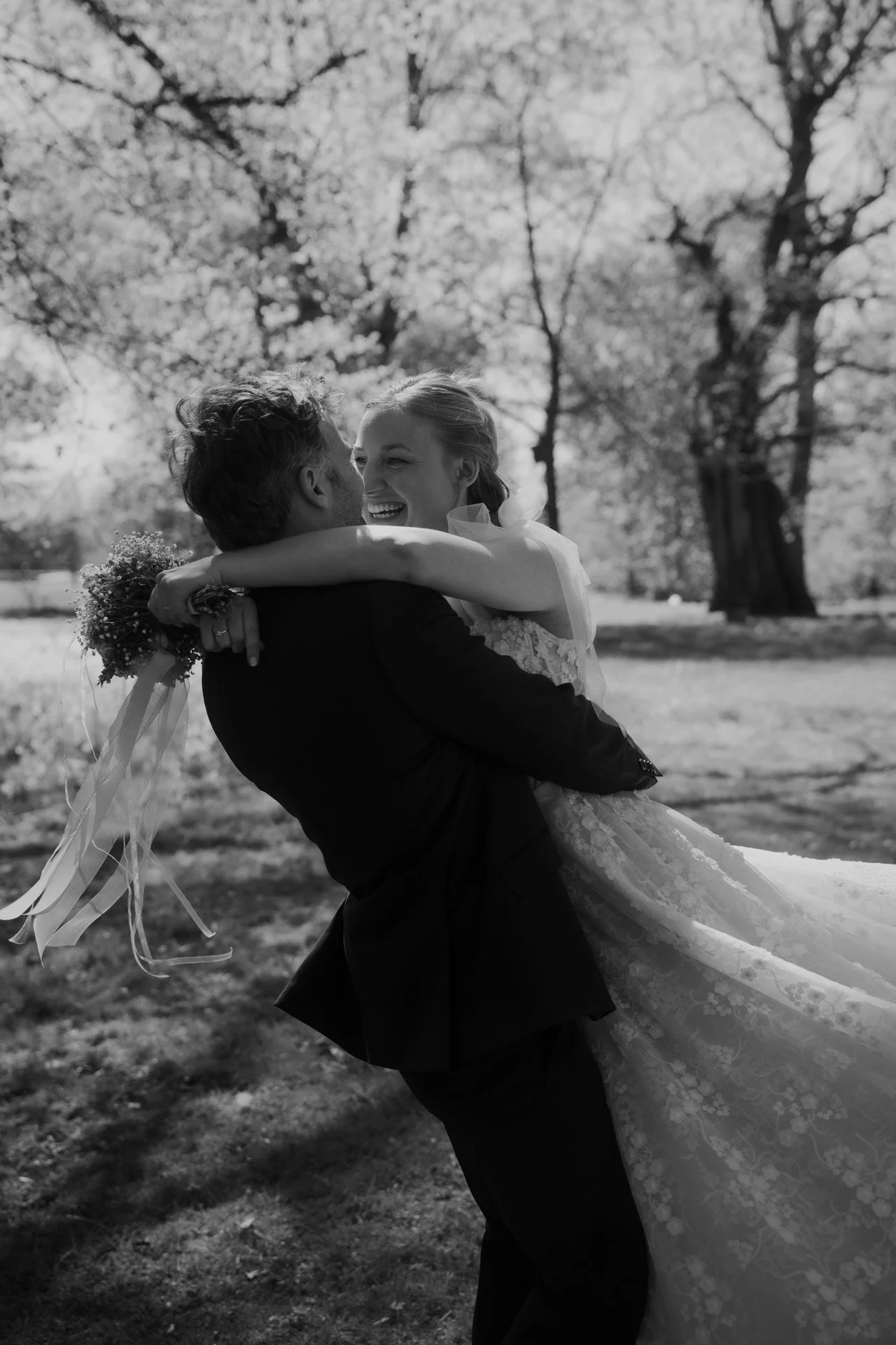 Ein Mann trägt eine Frau bei einer Hochzeit im Freien, umgeben von Bäumen, Lachen und Freude.
