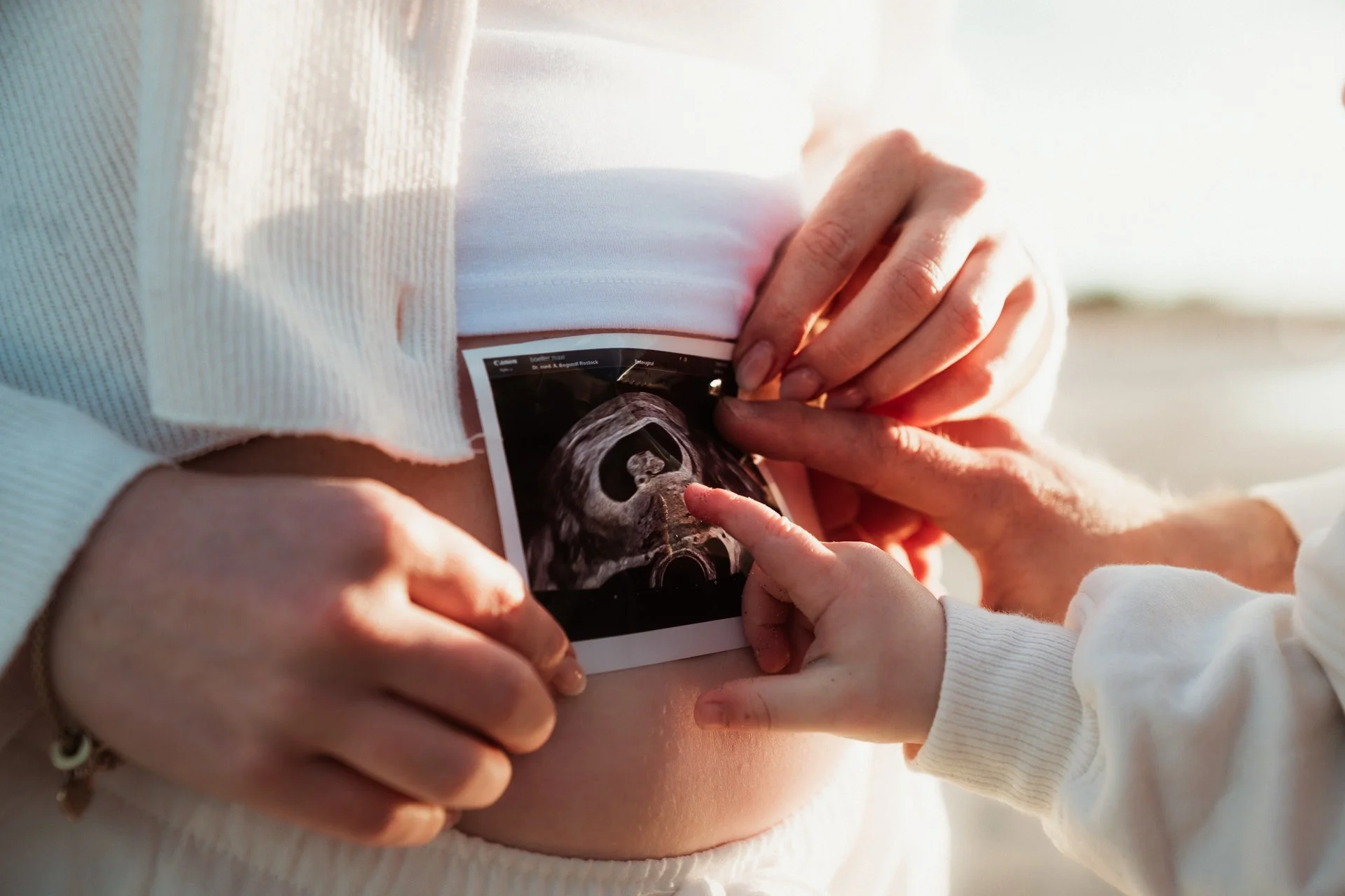 Eine Person zeigt eine Ultraschallaufnahme eines Babys in ihrem Bauch, während eine andere Person mit einem Finger darauf zeigt, vermutlich ein kleines Kind.