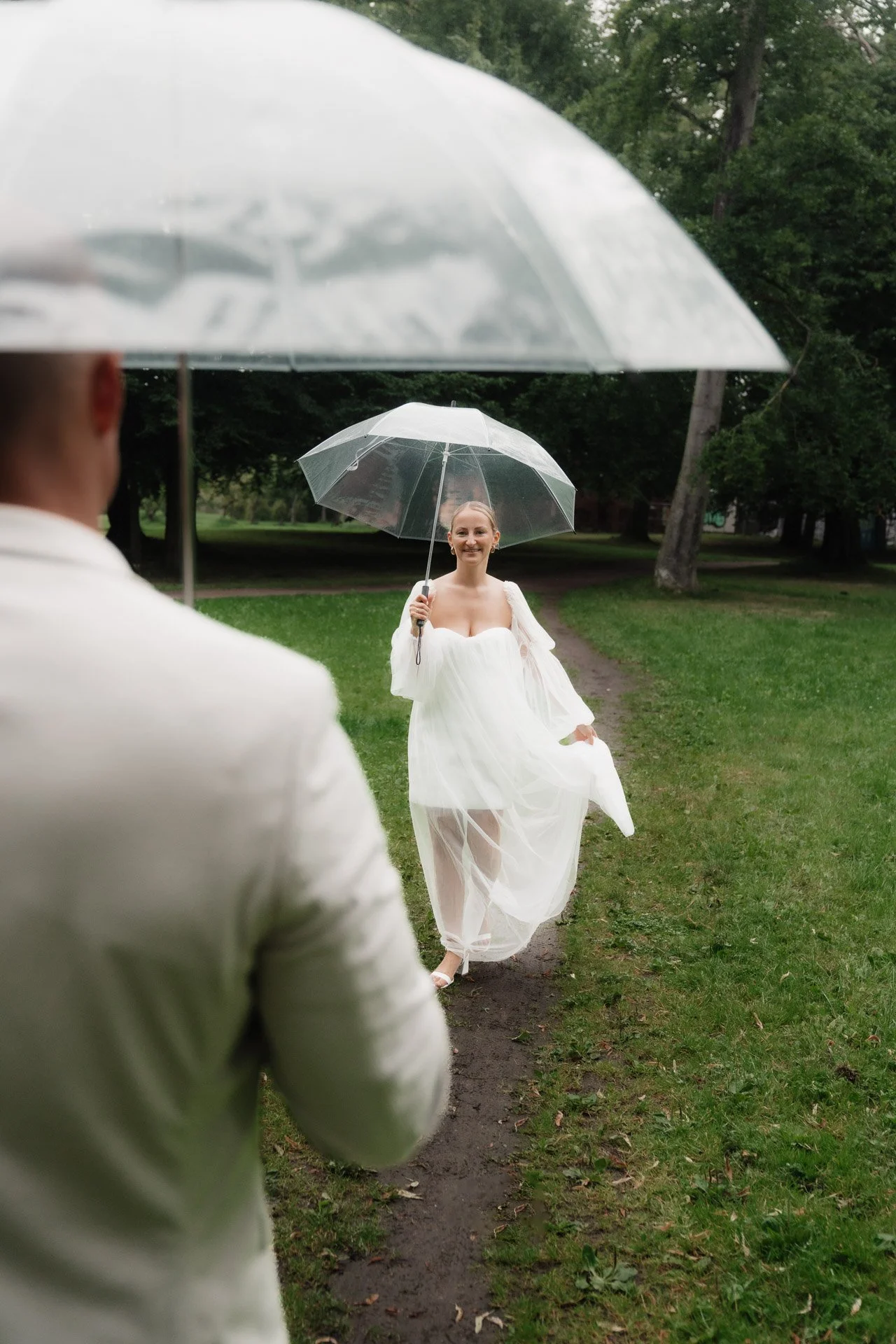Braut im weißen Kleid hält einen transparenten Regenschirm, während sie einem Mann mit einem weißen Anzug gegenübersteht, im Park bei Regen.