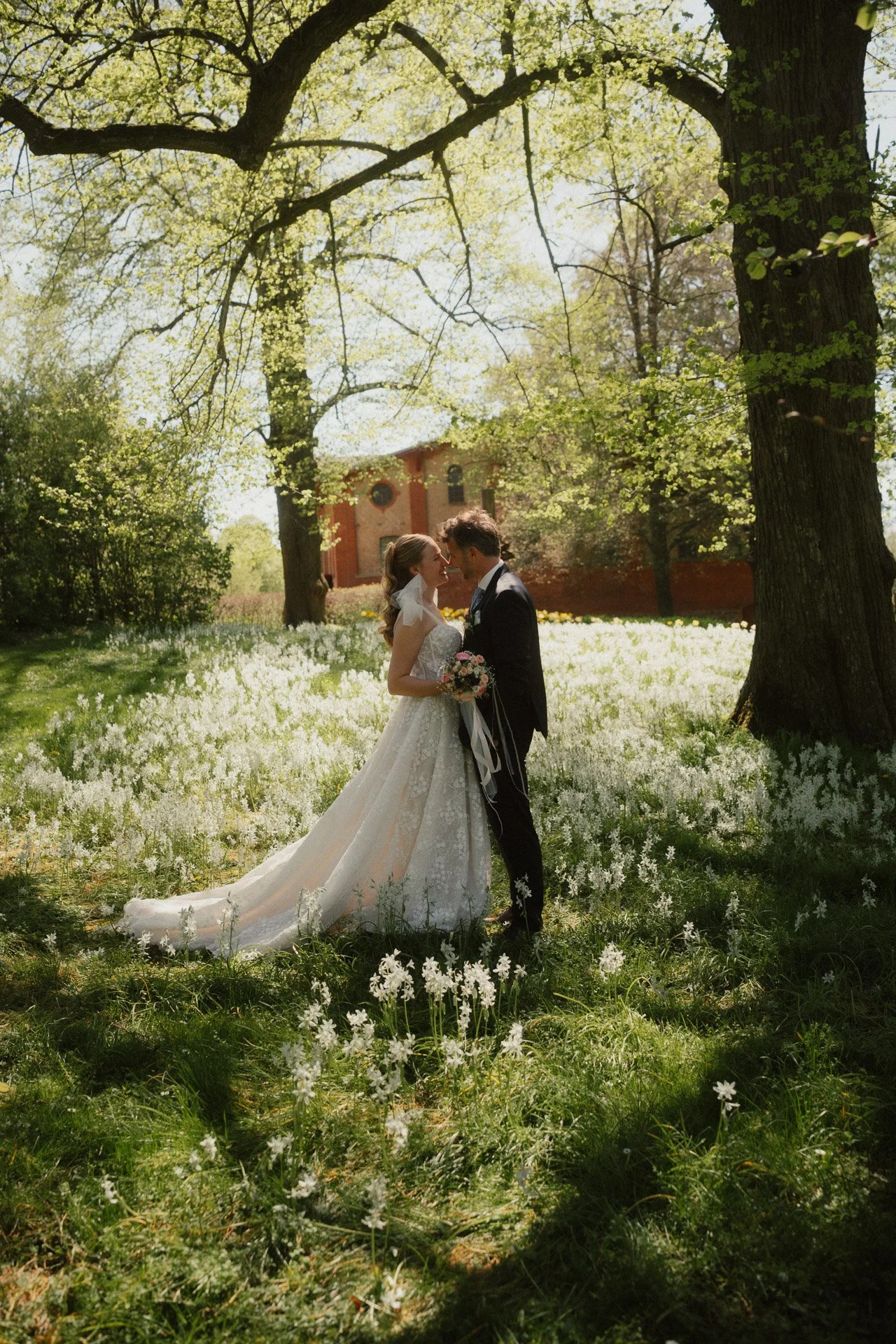 Ein Brautpaar steht sich in einem Garten mit weißen Blumen und zwischen Bäumen, im Hintergrund eine alte Kirche. Es wird wahrscheinlich geheiratet.