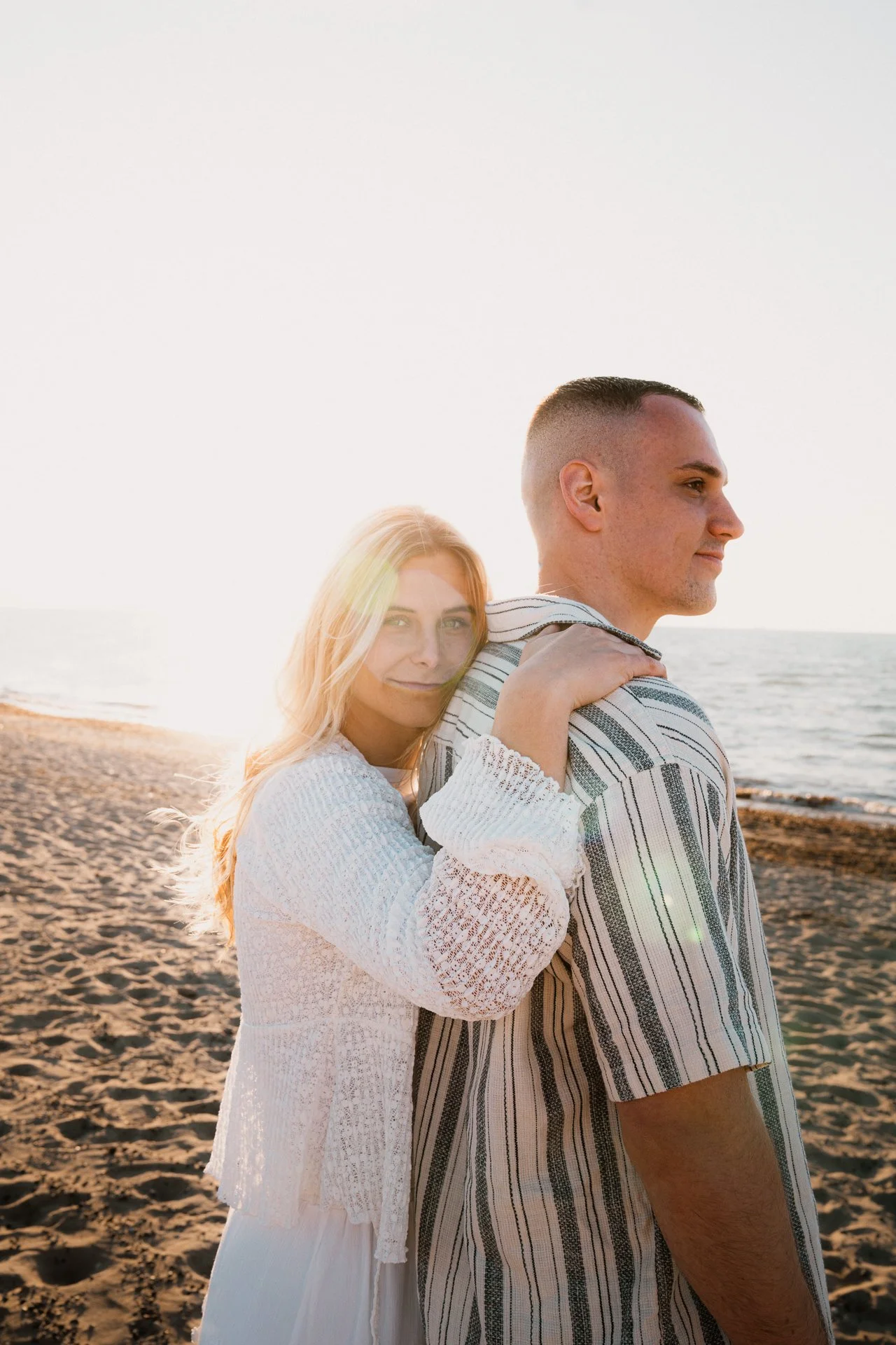 Ein Paar steht am Strand bei Sonnenuntergang, die Frau umarmt den Mann von hinten und schaut in die Kamera.
