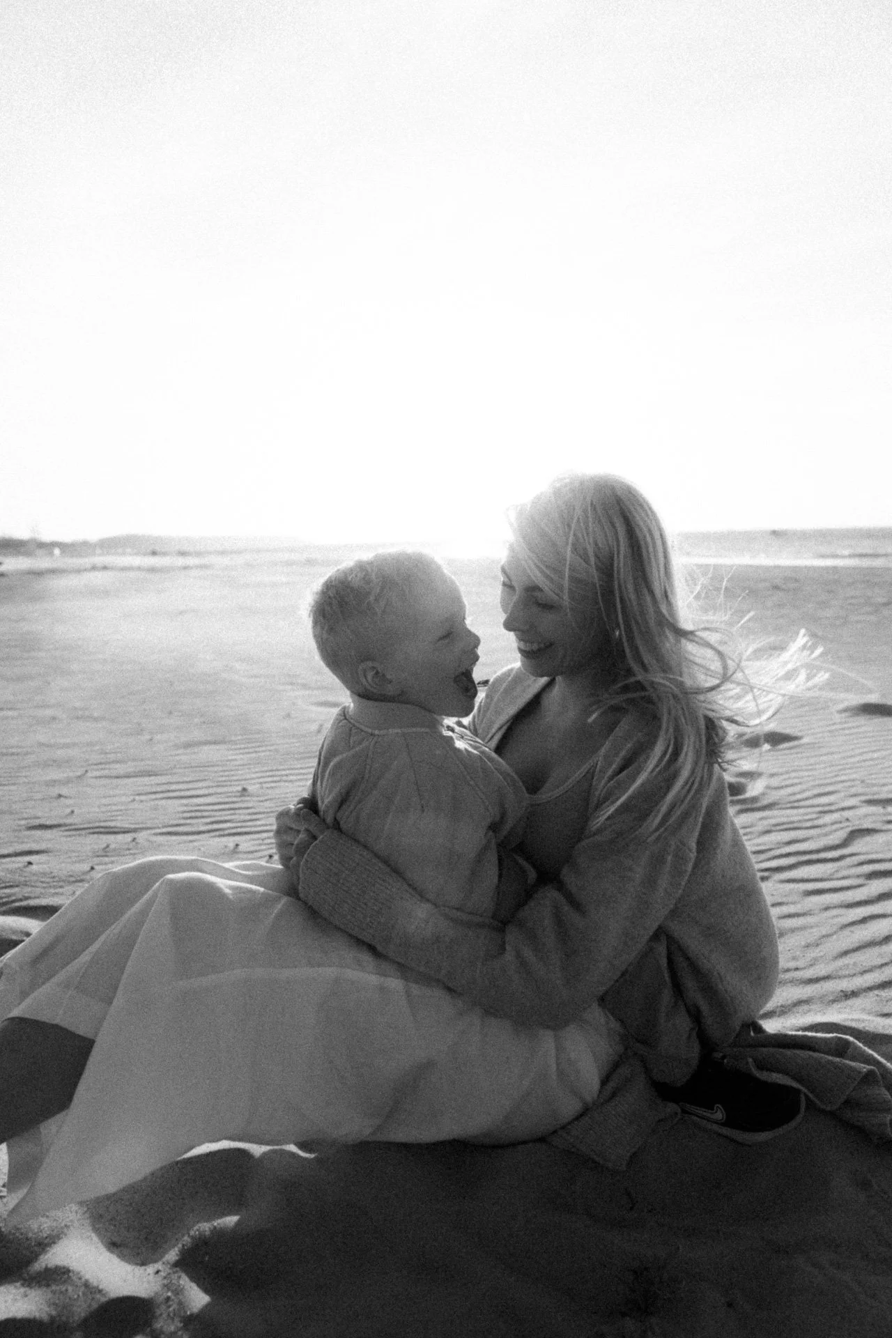 Eine lachende Frau und ein Kind sitzen am Strand, umgeben von Sand, bei Sonnenuntergang.
