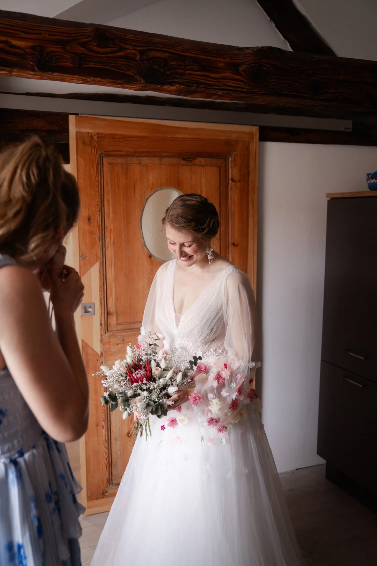 Eine lächelnde Braut in einem weißen Hochzeitskleid hält einen Blumenstrauß und steht neben einer Freundin, die vor Freude lacht.