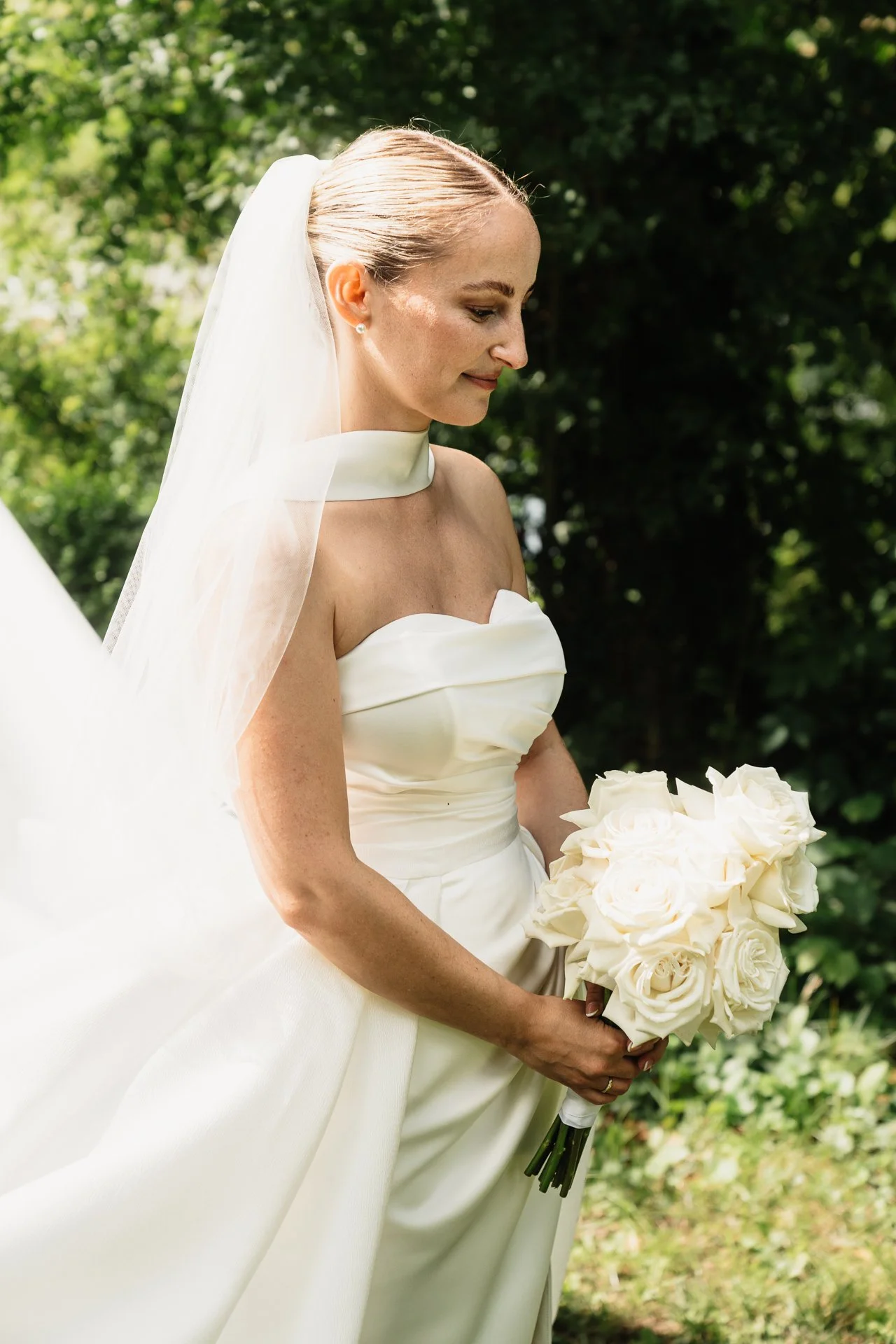 Braut in einem weißen Hochzeitskleid mit Schleier hält einen Blumenstrauß aus weißen Rosen im Freien an einem sonnigen Tag.
