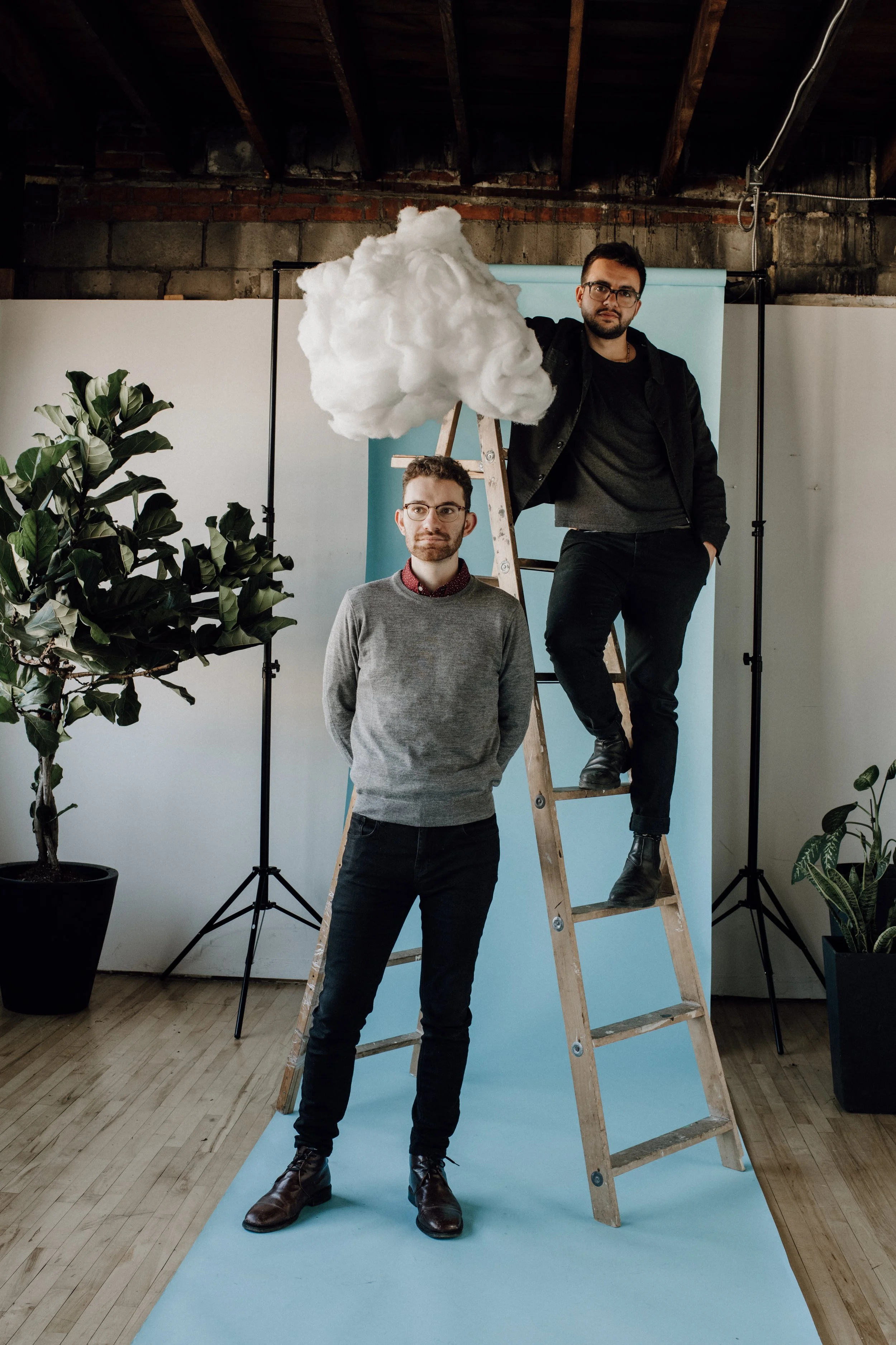 Two men in a photography studio, one standing on the floor and the other sitting on a ladder, holding a cloud-shaped prop over his head. There are plants and photography equipment around.