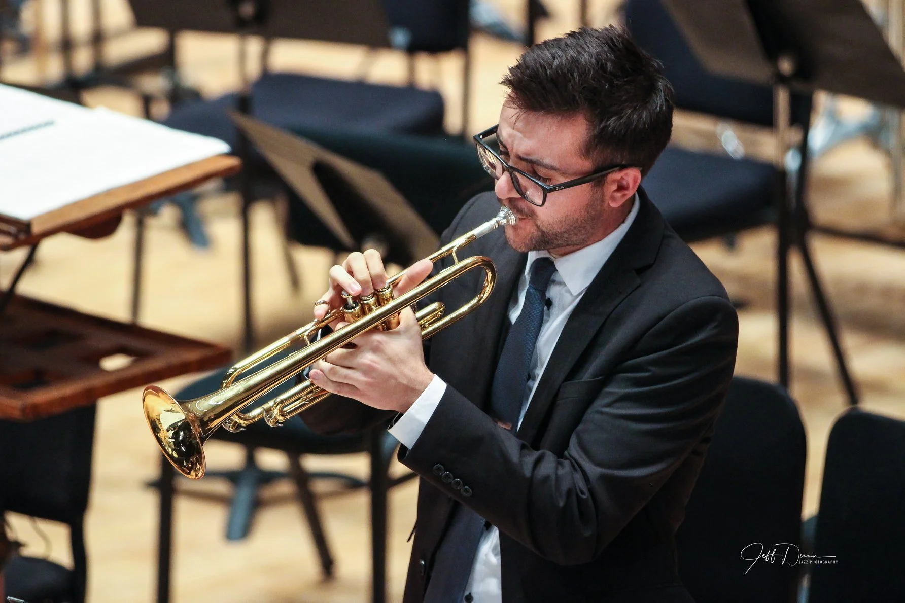 A man in a black suit, white shirt, and tie playing a trumpet during a performance, with music stands and chairs in the background.