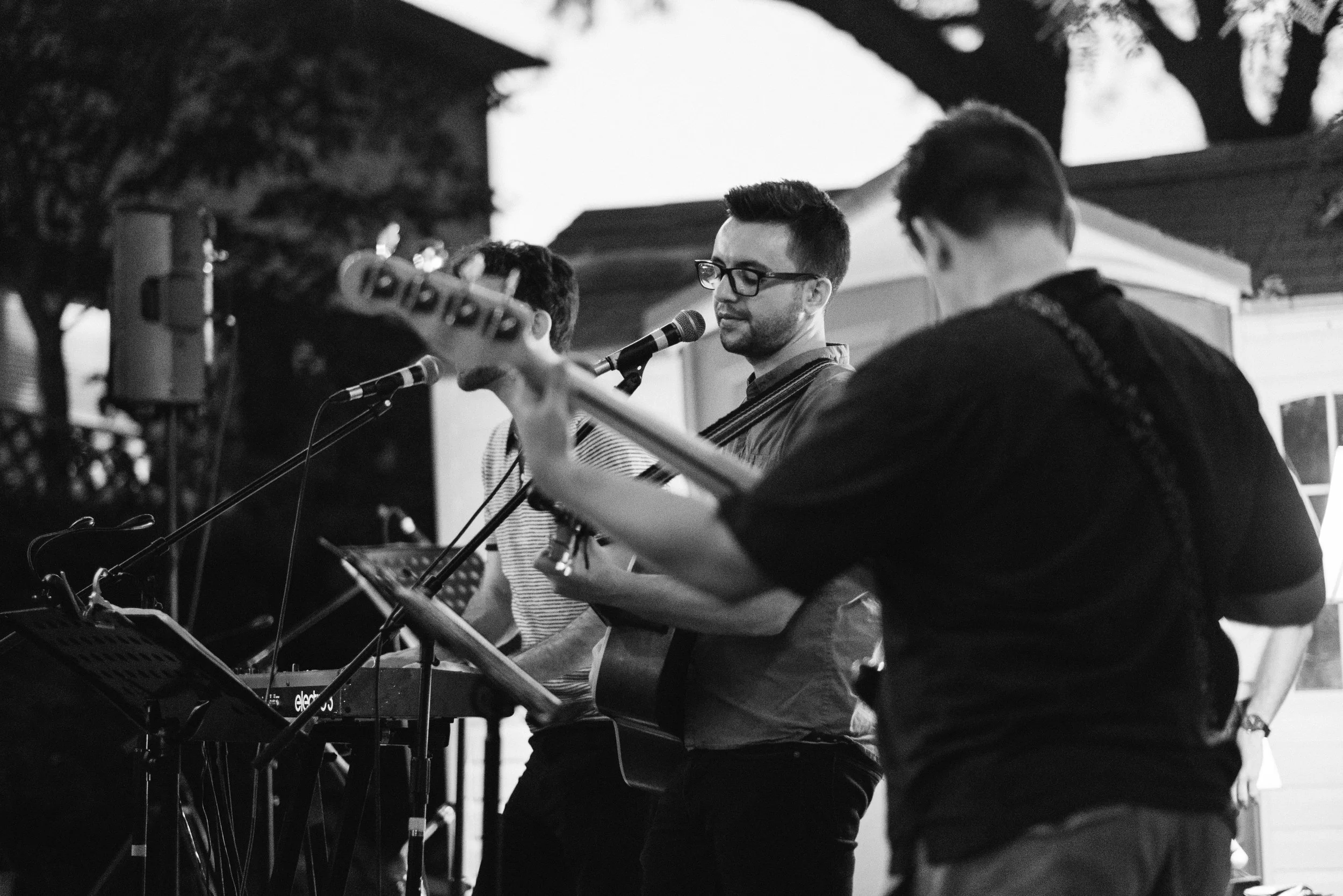 A group of musicians performing outdoors, including a man with glasses singing into a microphone, a guitarist in the foreground, and other band members playing instruments, with trees and tents in the background.