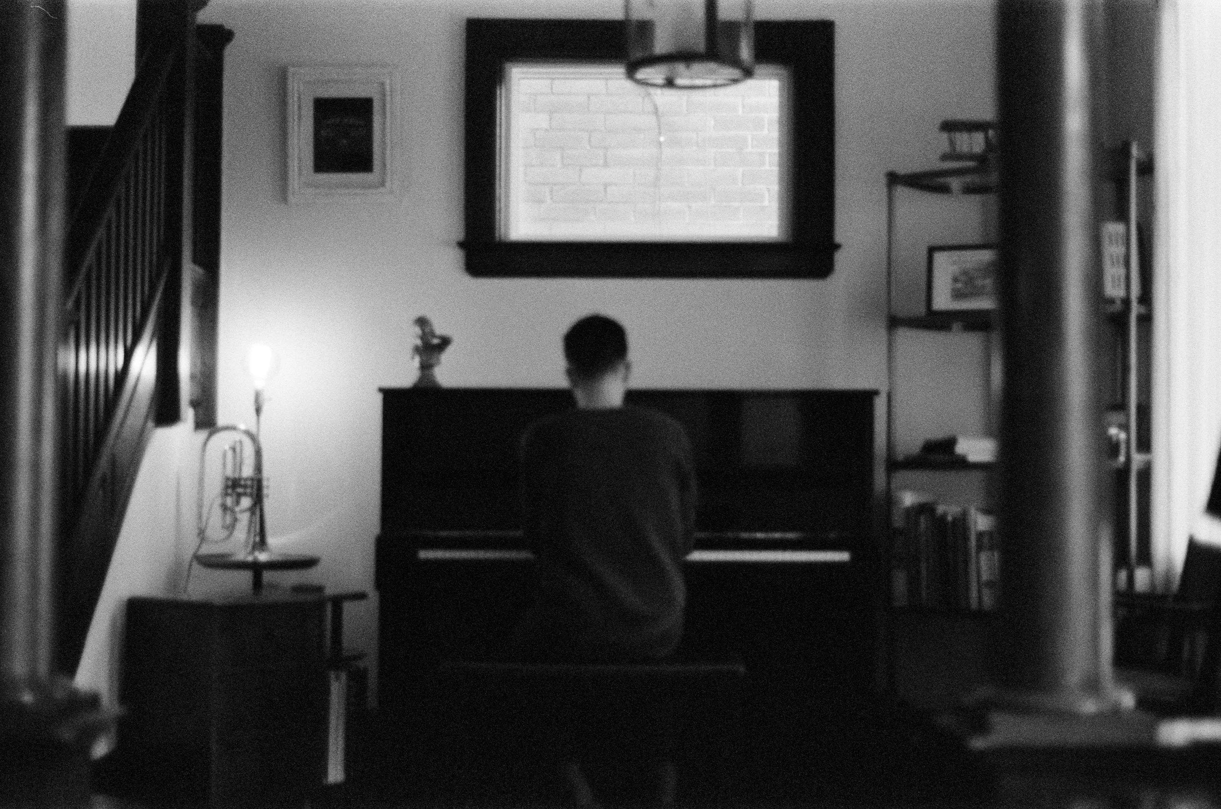 A person sitting at a piano in a living room, seen from behind, with a window and bookshelf in the background.