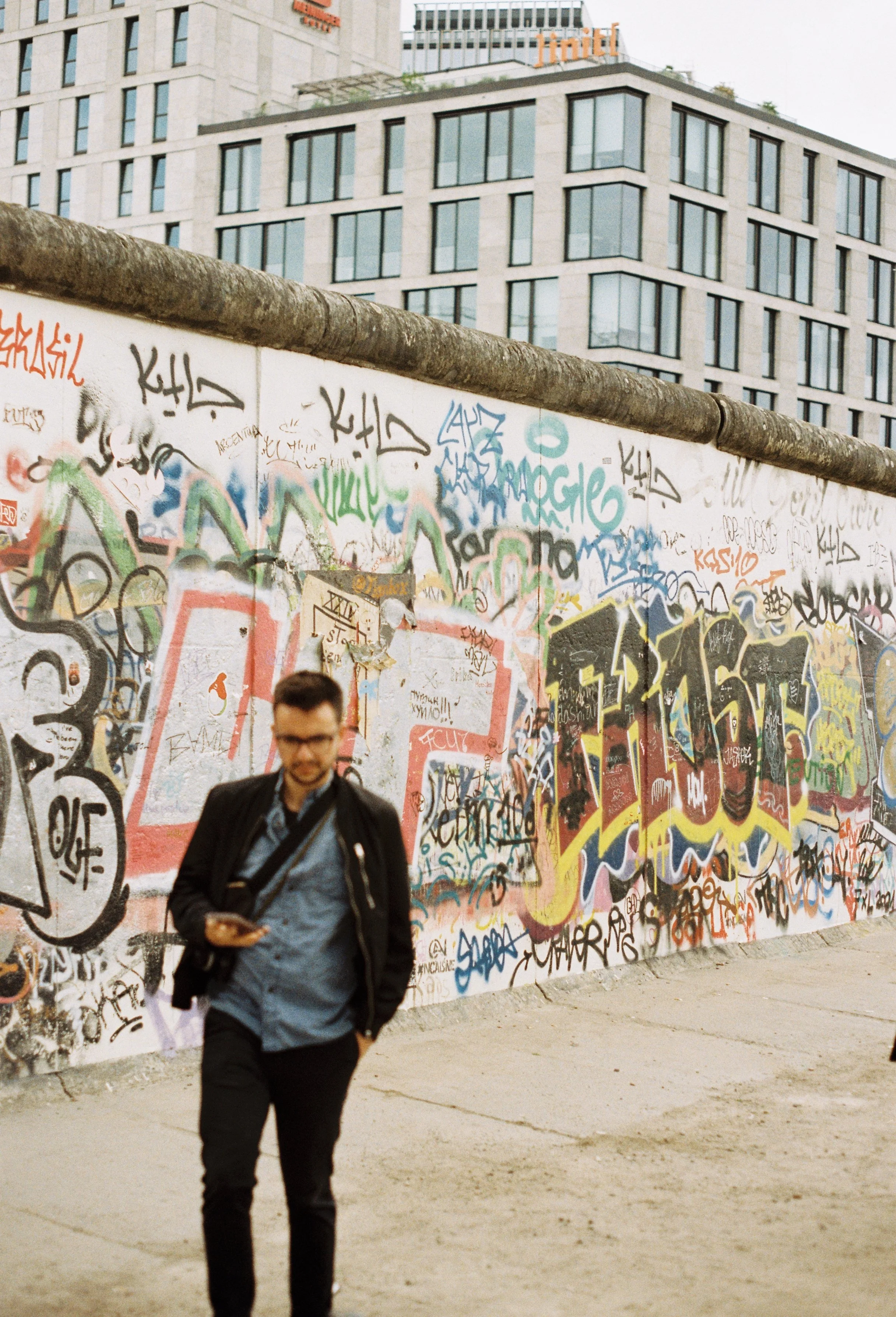 A man with glasses, wearing a black jacket and blue shirt, walking along a graffiti-covered section of the Berlin Wall. 