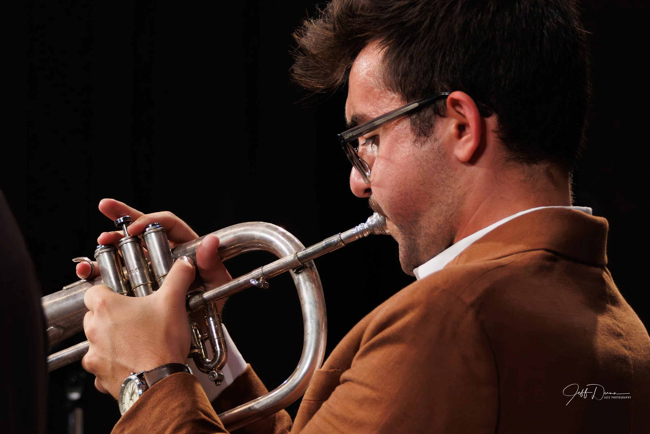 A man playing a trumpet, wearing glasses and a brown jacket, with a black background.