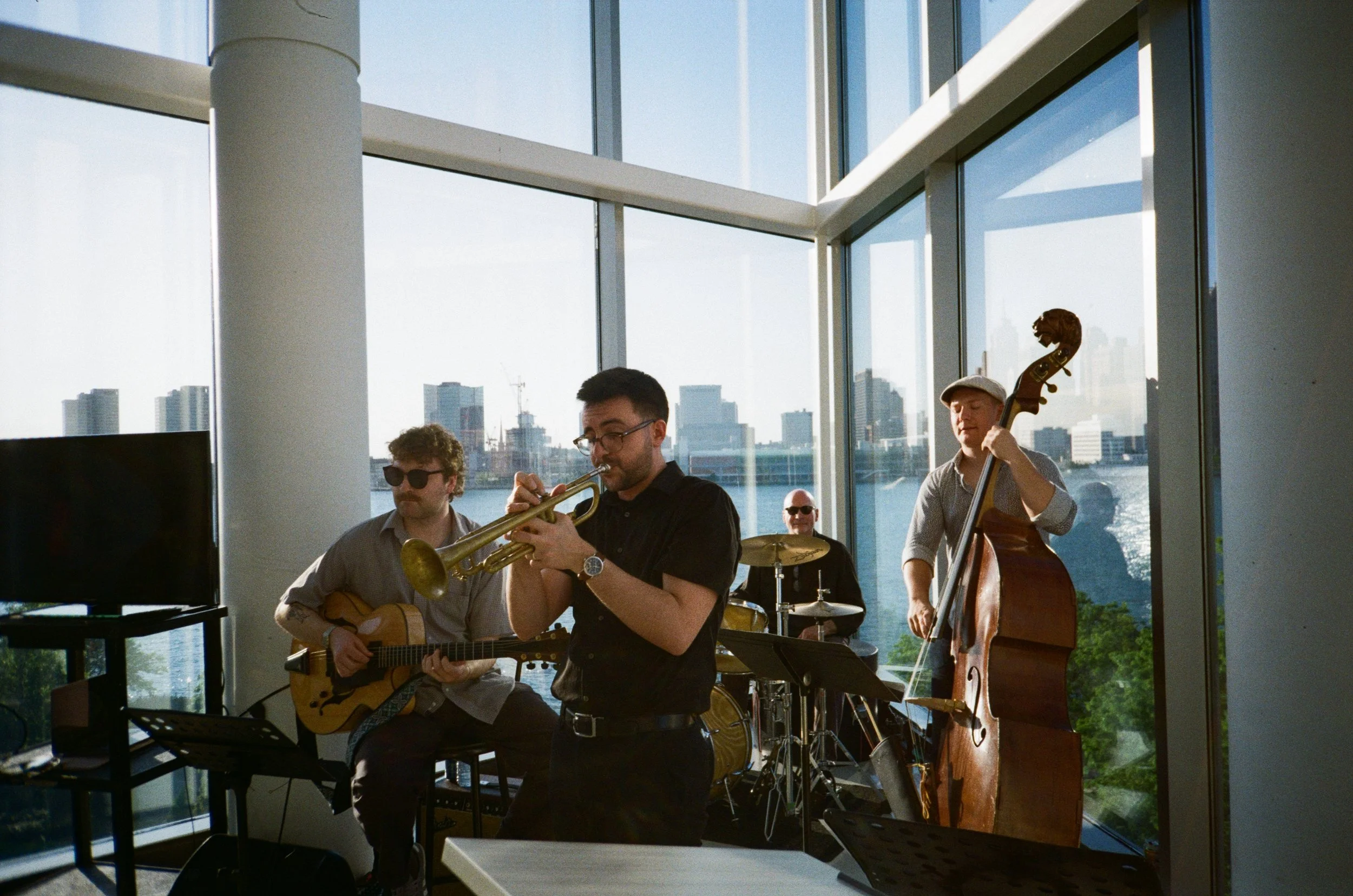 A four-piece jazz band performing in a modern, glass-walled room with a cityscape and river view in the background. The band includes a trumpeter, guitarist, drummer, and double bass player.