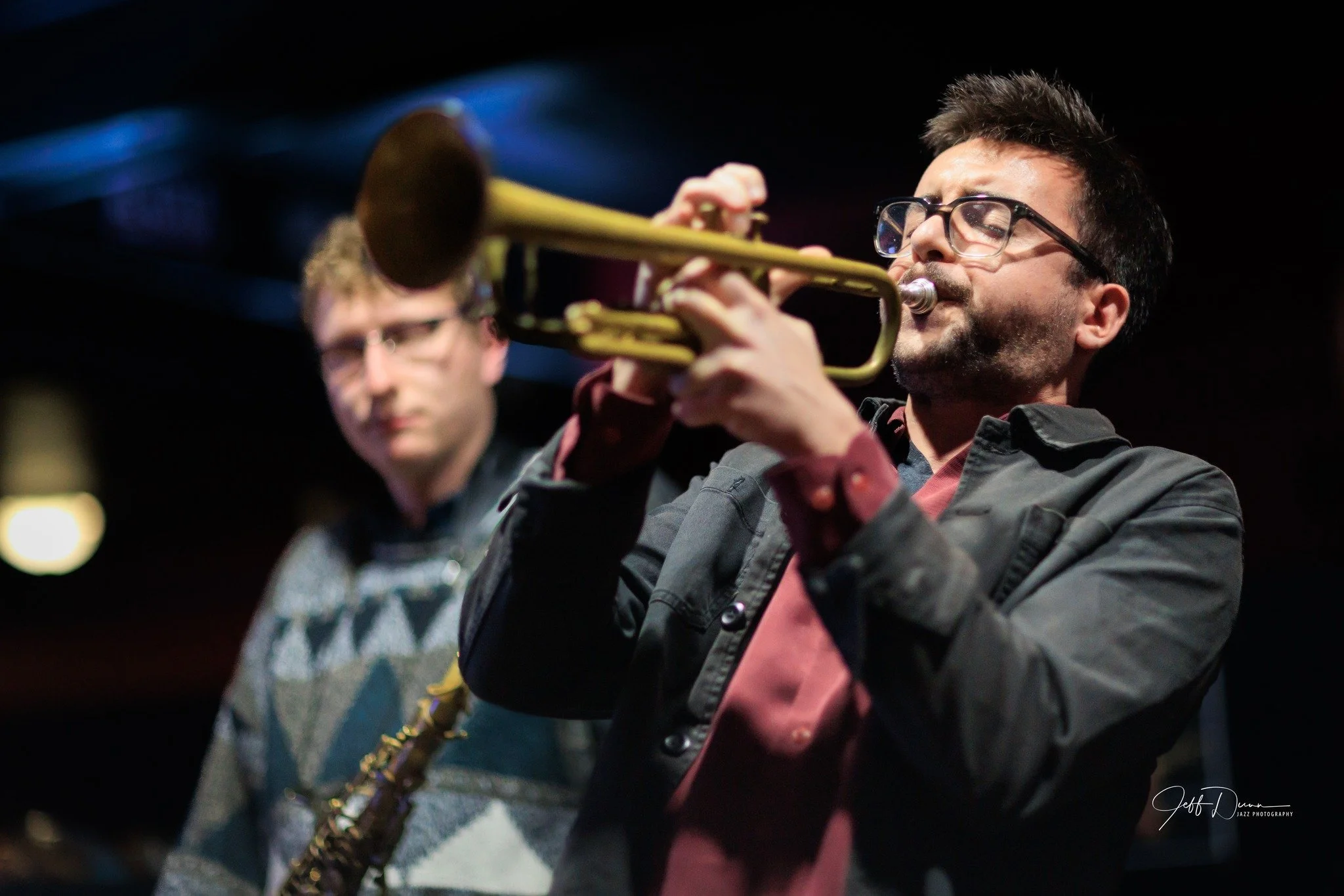 Two musicians performing, one playing the trumpet and the other on the saxophone, in a dimly lit setting.