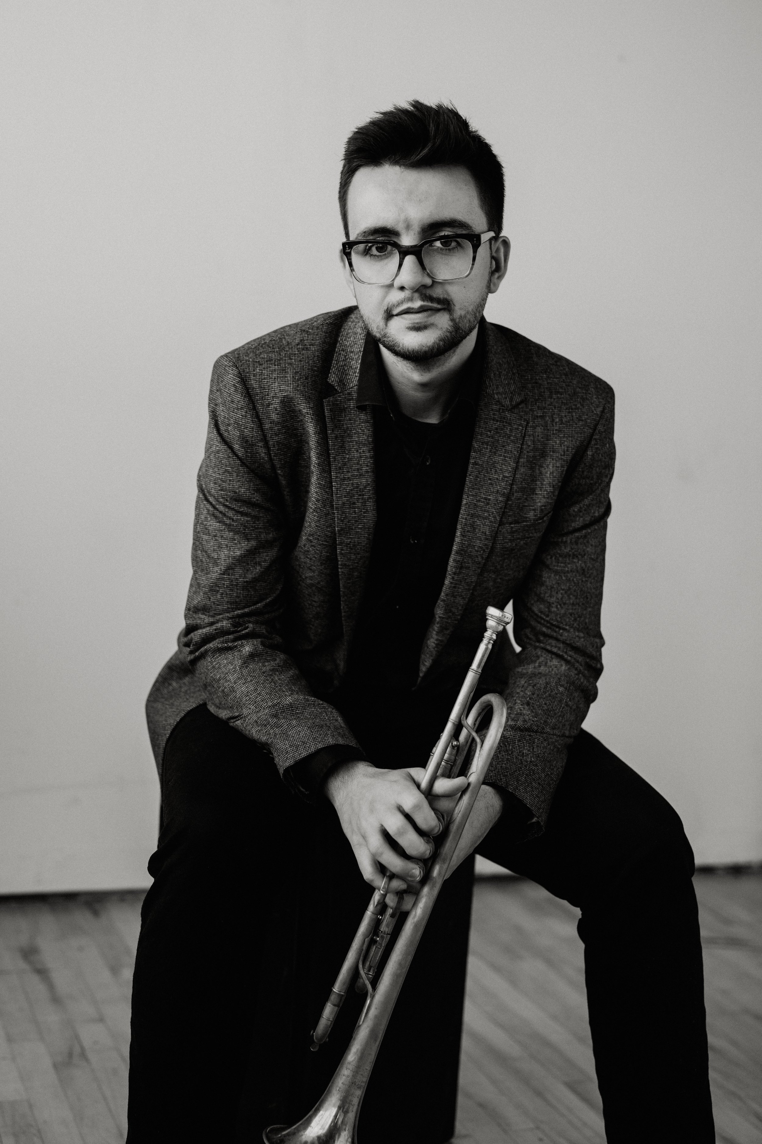 Black and white photo of Austin Di Pietro with glasses holding a trumpet, sitting on a stool against a plain wall.