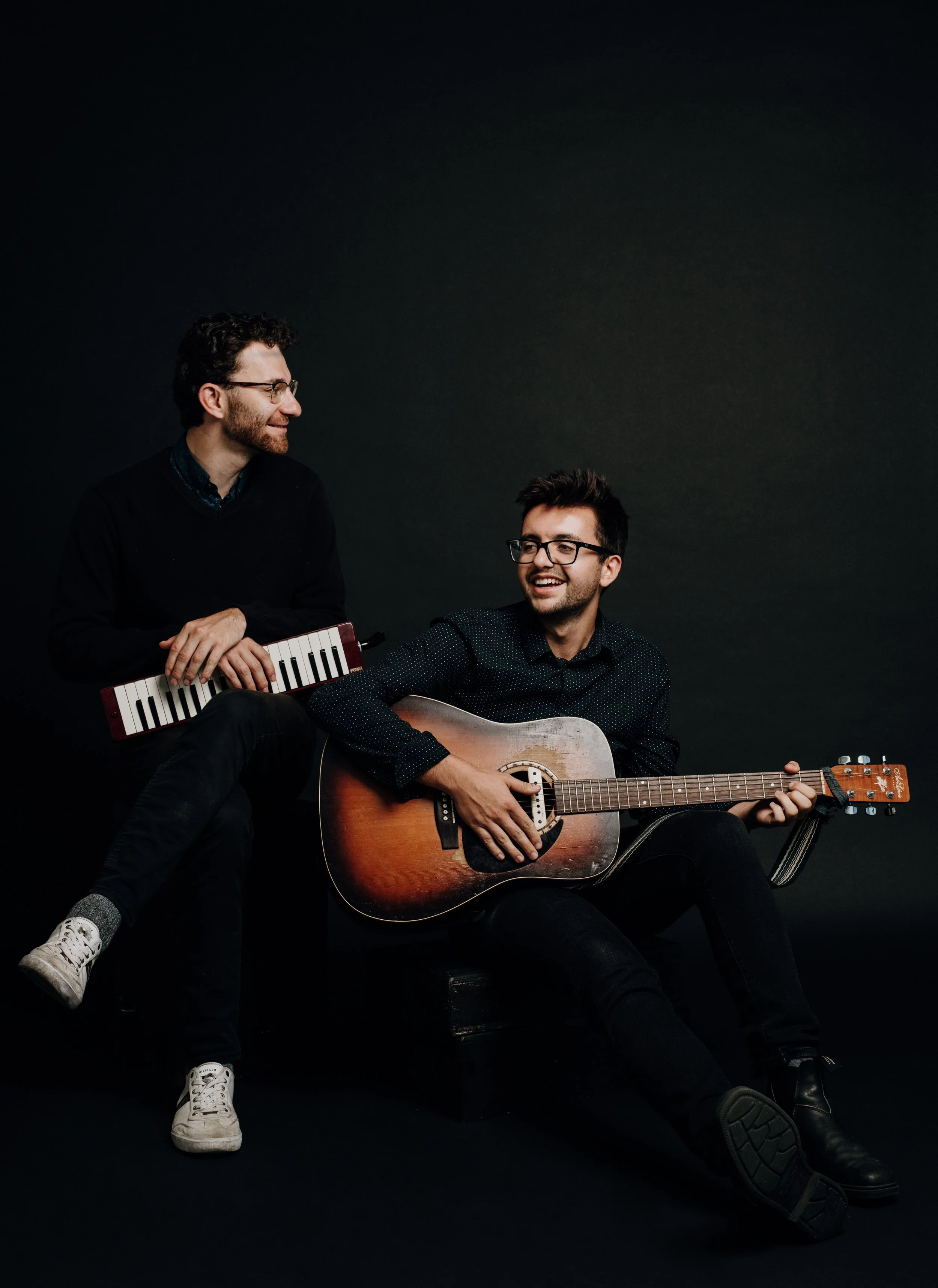 Two men with glasses (The Bishop Boys), one sitting on a black stool playing an acoustic guitar, and the other standing beside him holding a small keyboard, both smiling and looking at each other against a dark background.