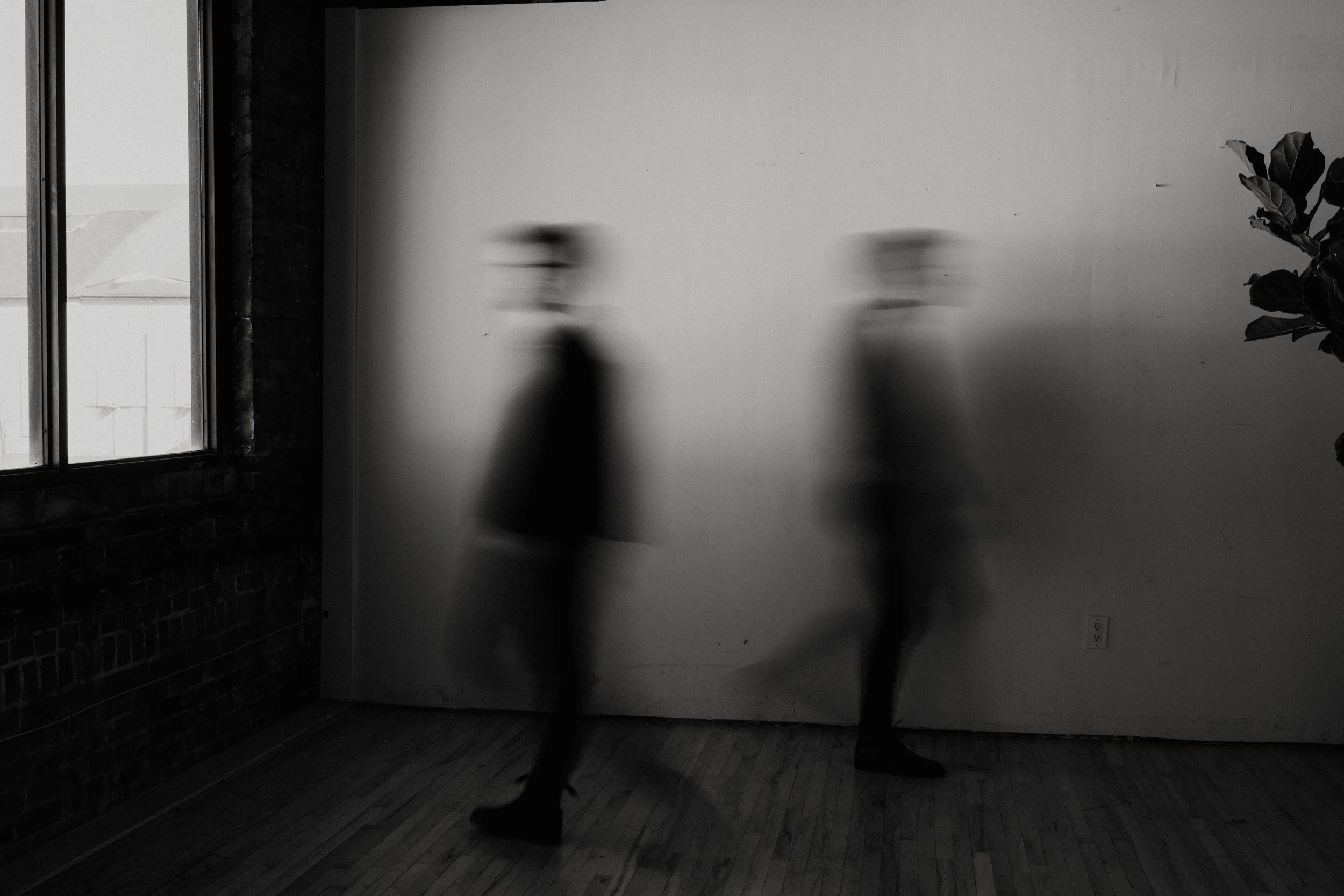 Two blurred individuals walking past each other against a white wall in a room with a brick wall and large window.