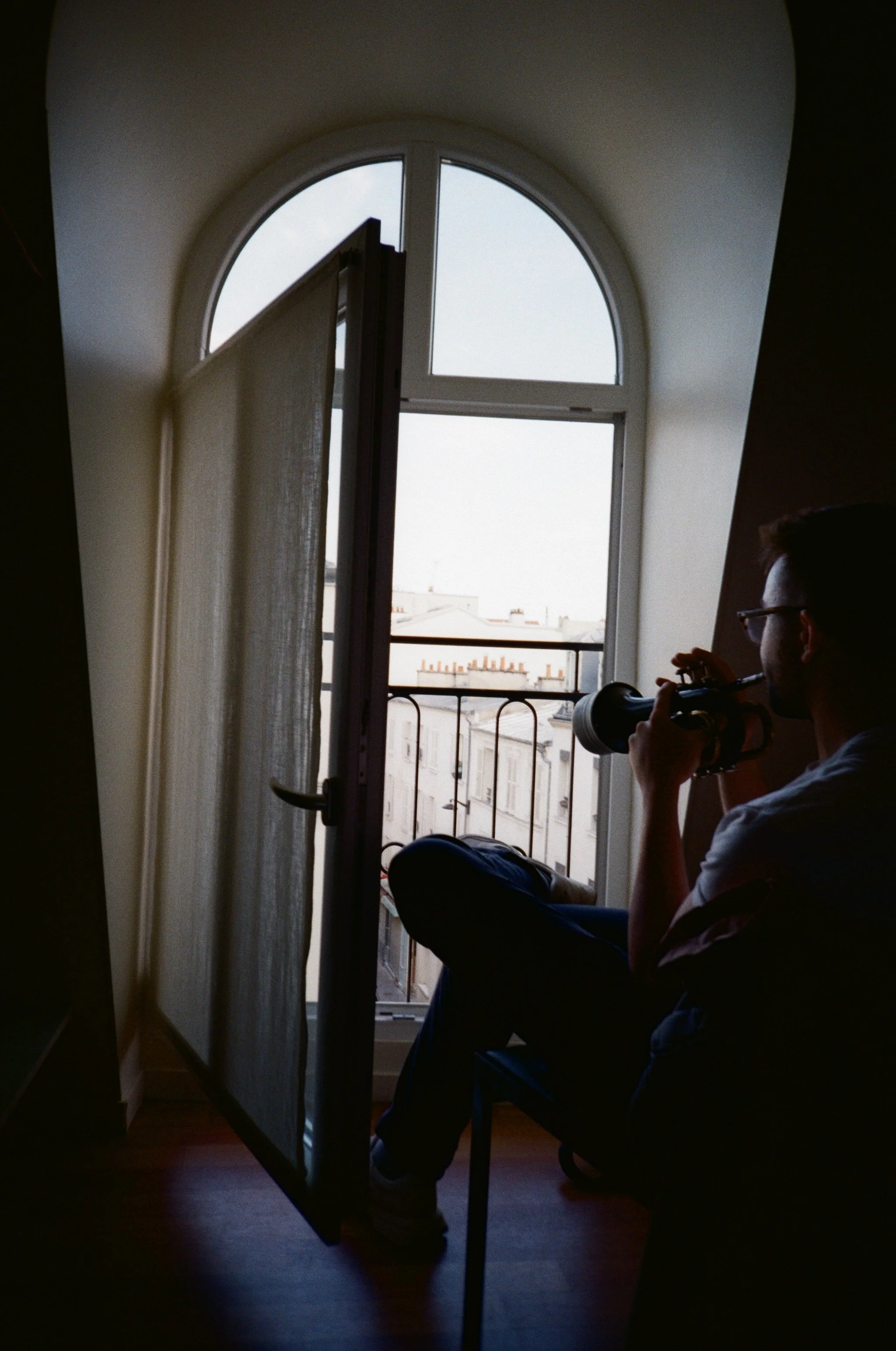 A person sitting on a chair near an open window, playing trumpet, inside a room during daylight.