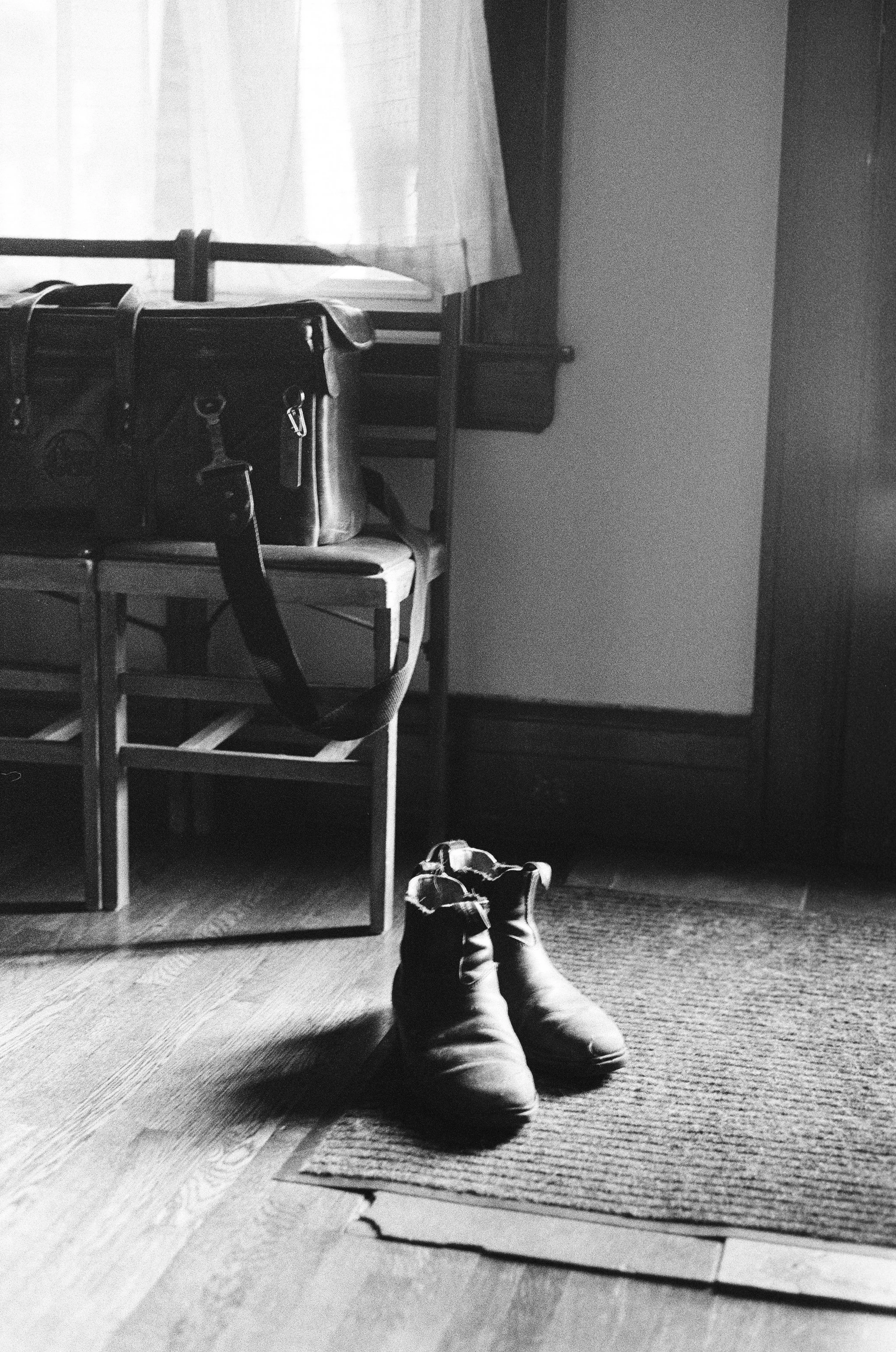 A black and white photo of a pair of boots on a rug near a door, with a chair, a bag, and a window with curtains in the background.