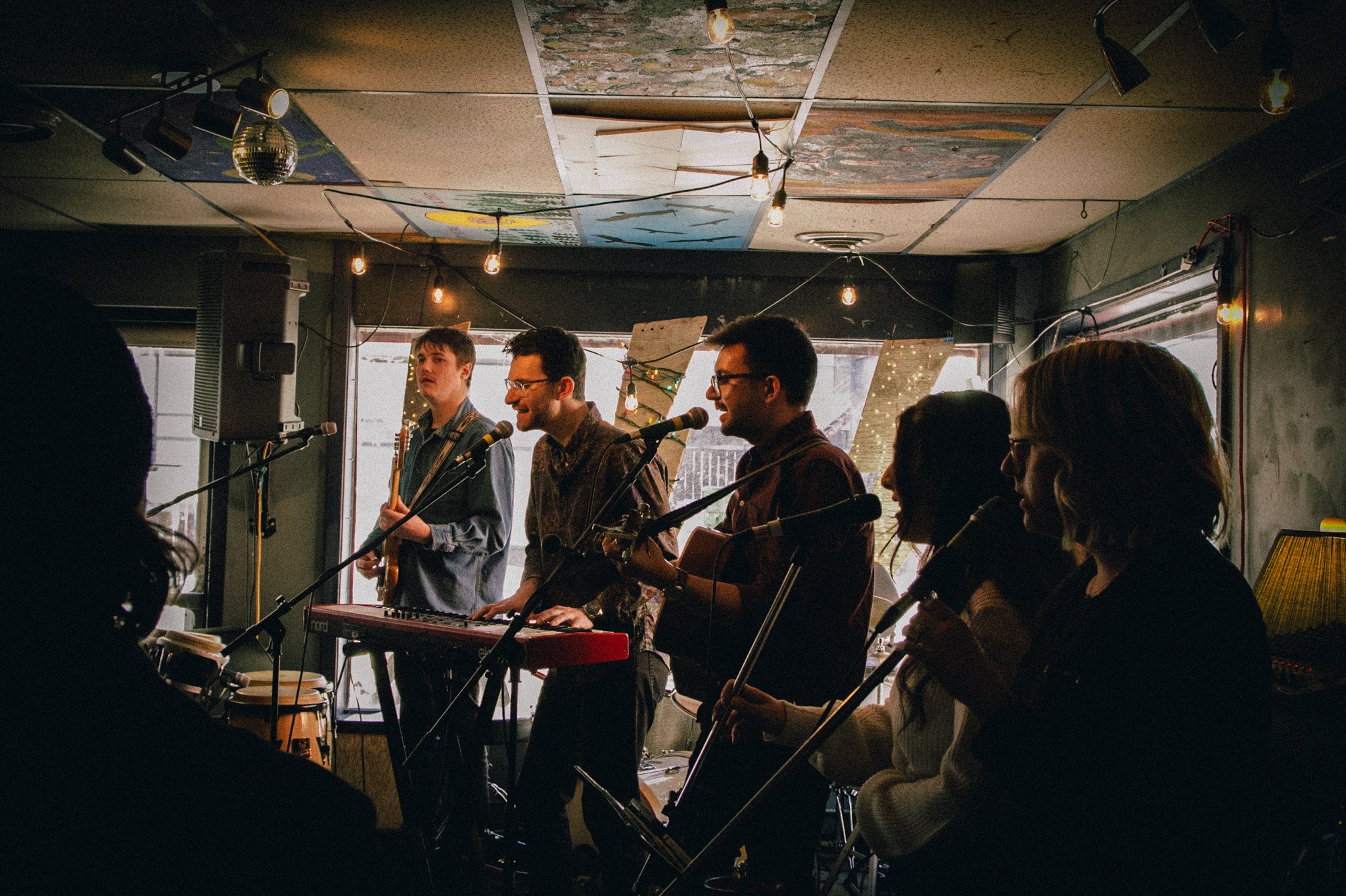 A group of six musicians performing live in an intimate, dimly-lit venue with string lights, a ceiling decorated with artwork, and a large window in the background, featuring a keyboard, microphones, and musical instruments.