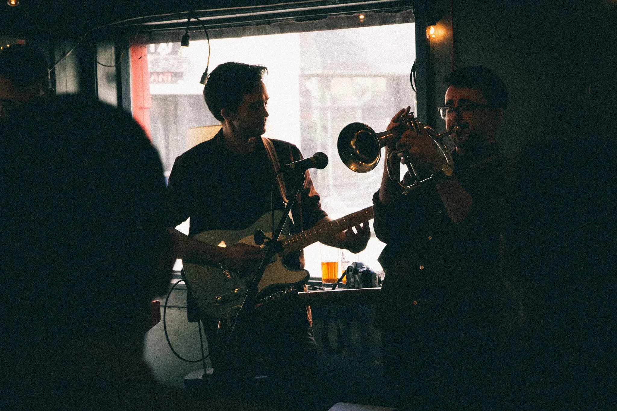 Musicians performing in a dimly lit indoor setting with one playing guitar and the other playing trumpet, near a window with natural light.