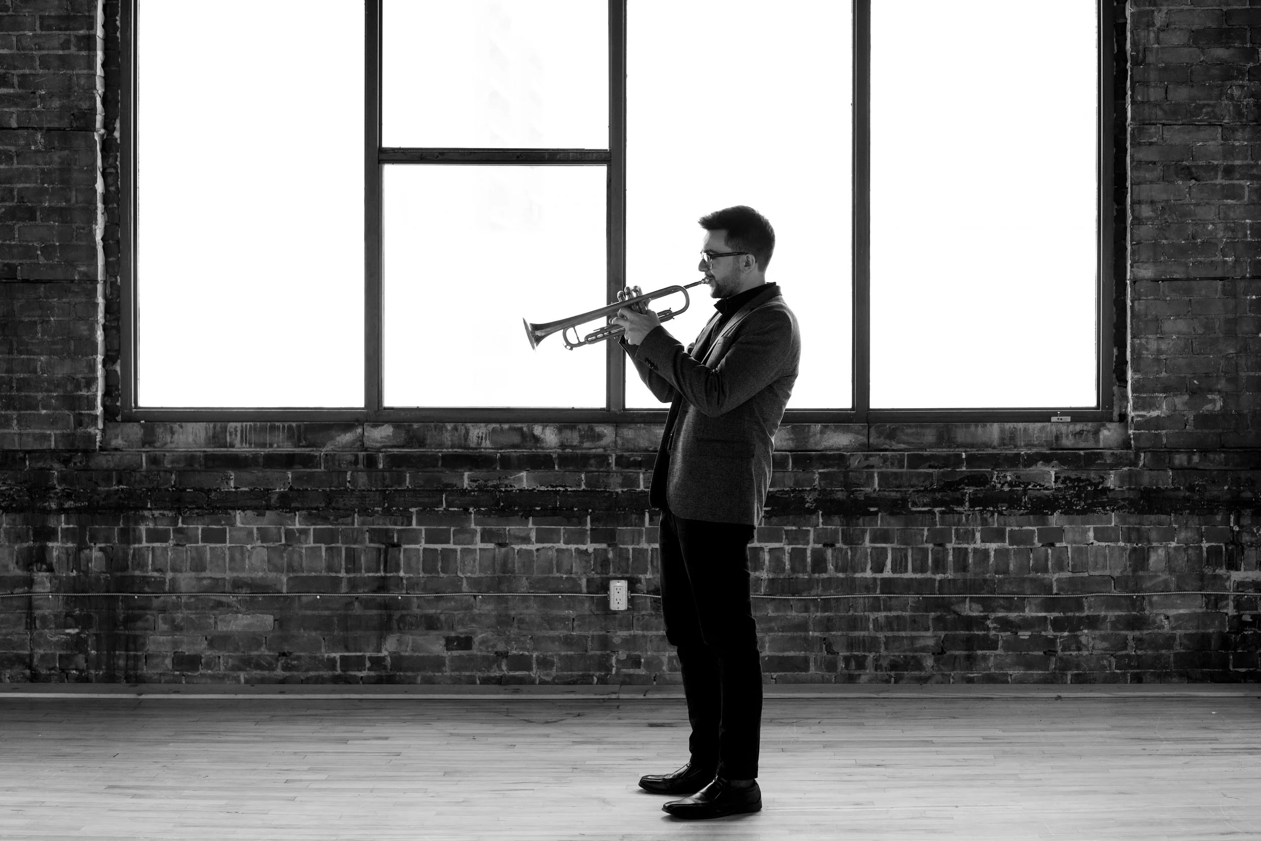 A man in a blazer and glasses playing a trumpet in a room with large windows and brick walls.