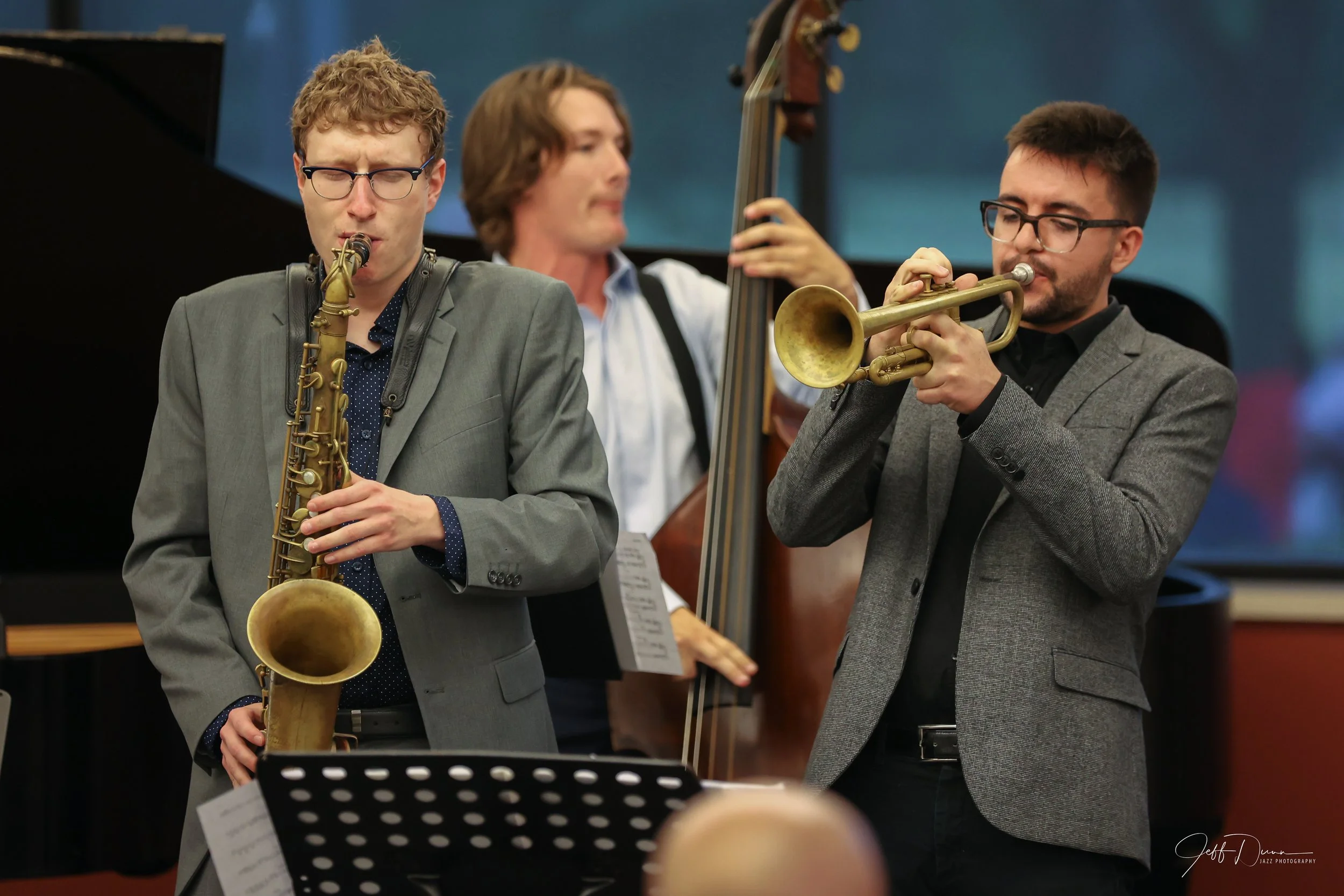Three male jazz musicians performing, with a saxophone and trumpet players in the foreground and a double bassist in the background.